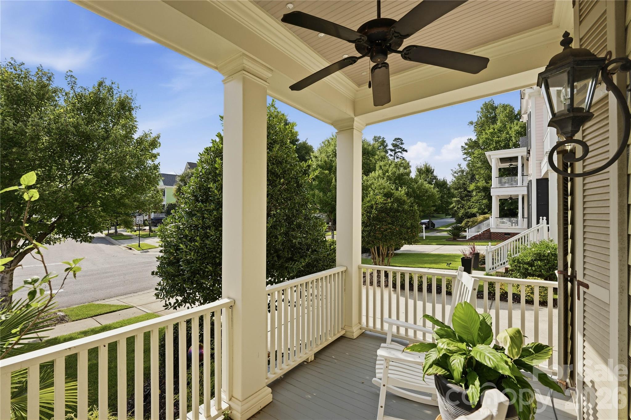 Covered front porch with white rocking chairs, potted plants, and a view of the leafy street.