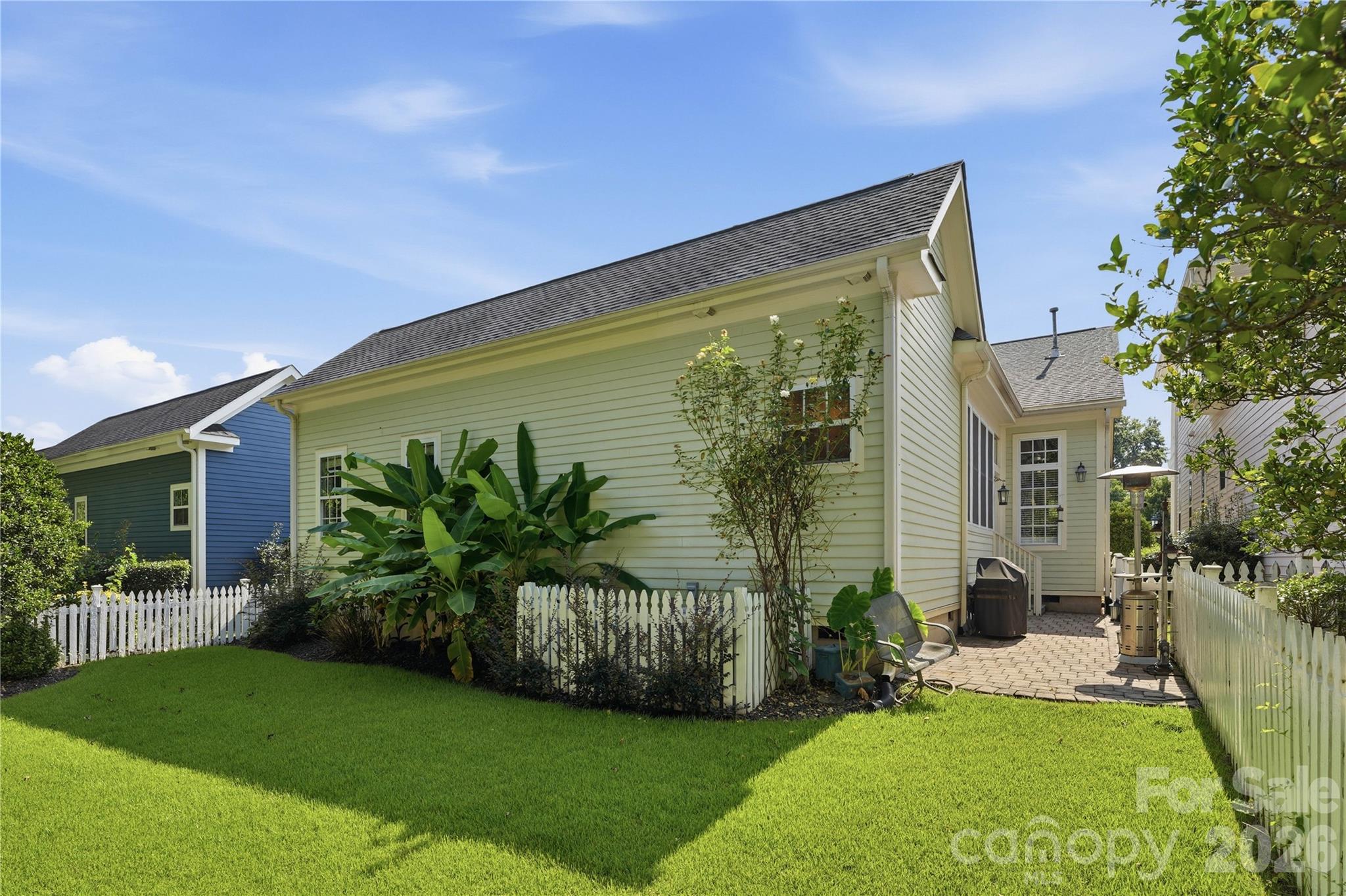Pale yellow home beside a fenced patio with a paved seating area and lush landscaping.