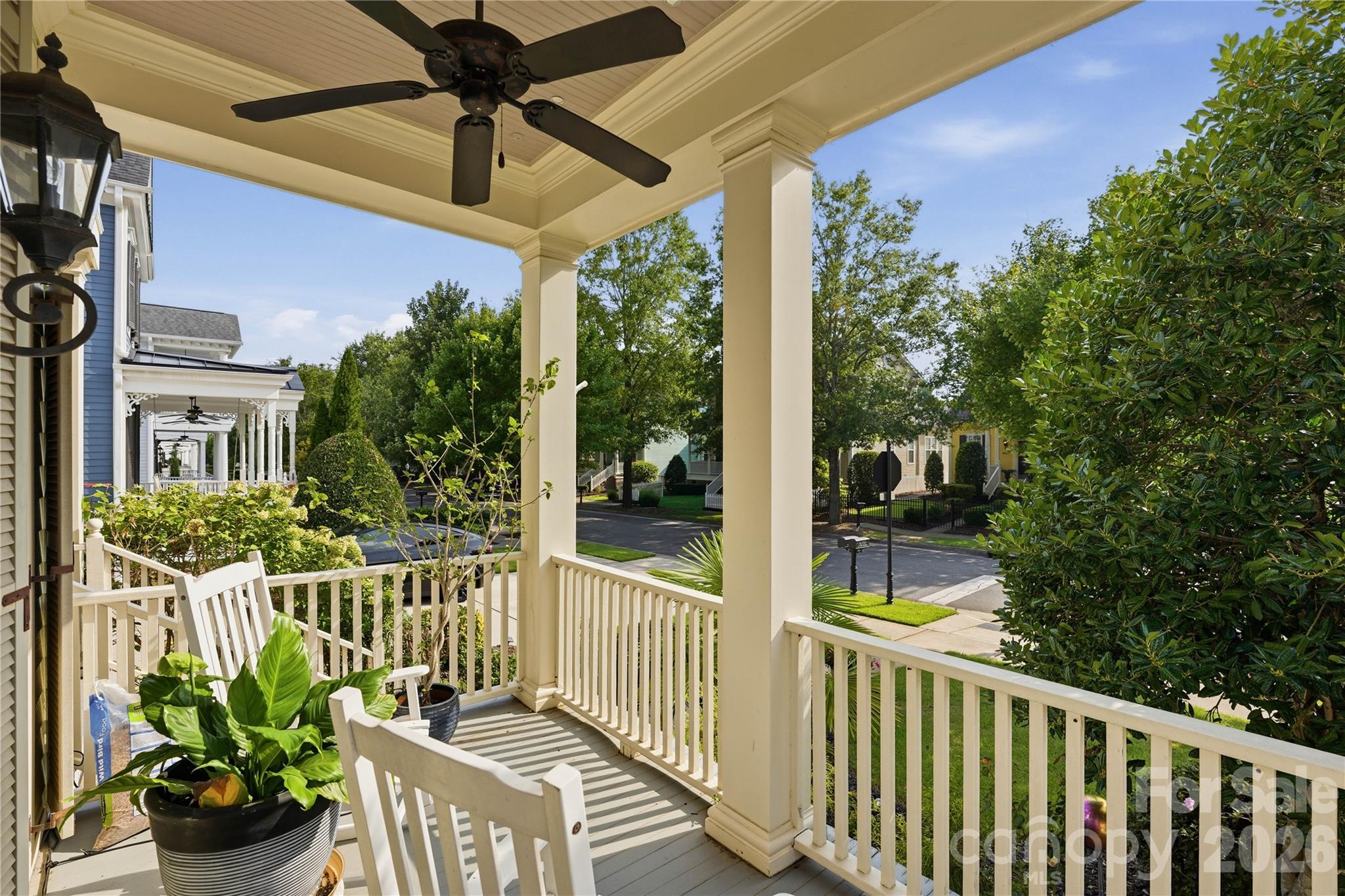 Covered front porch with cream columns, white railings, a ceiling fan, and potted plants overlooking a residential street.