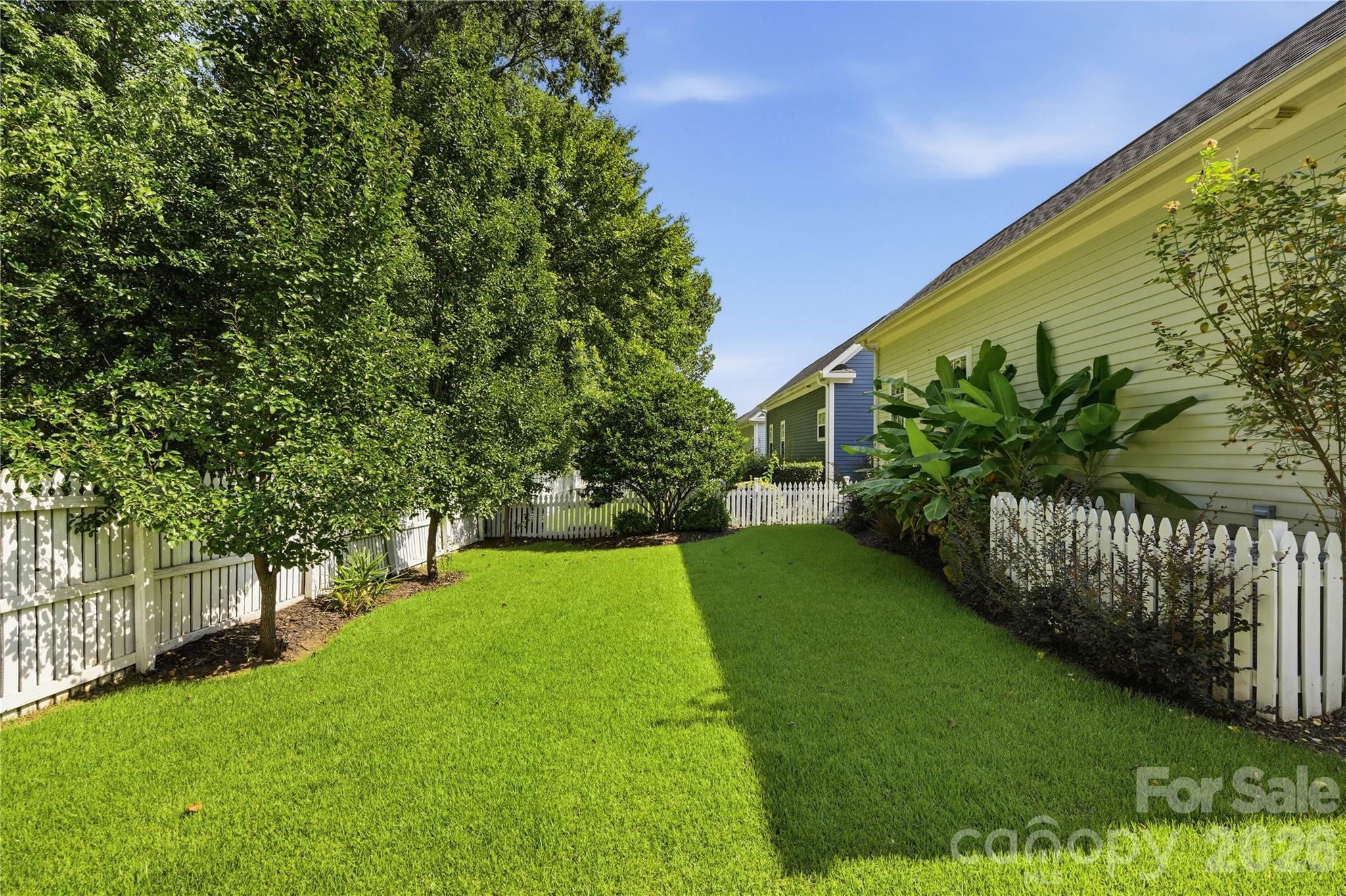Side yard with a broad lawn, white picket fences, mature trees, and leafy shrubs beside the house.