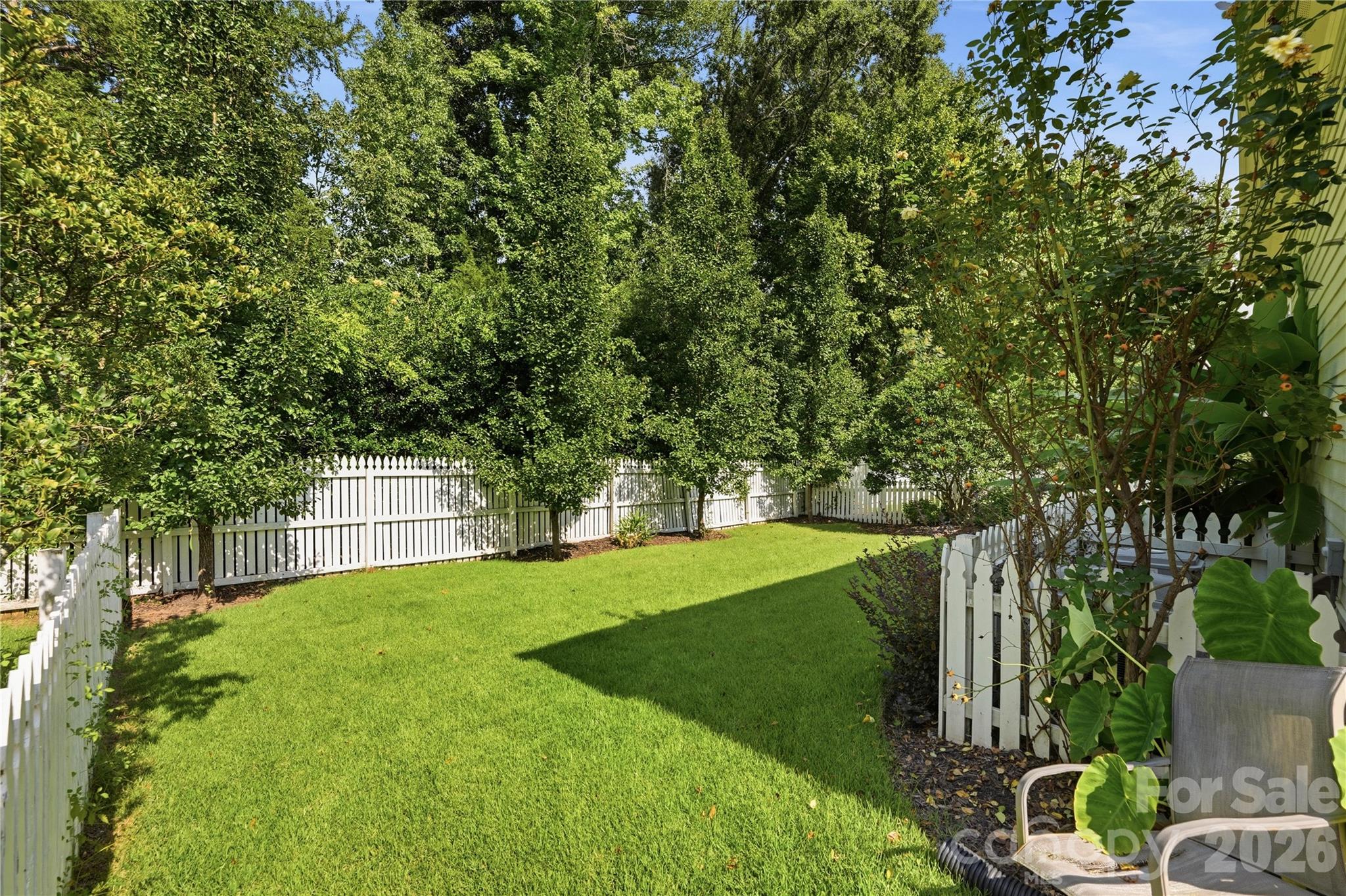 Fenced backyard with a green lawn, mature trees, white picket fence, and patio chairs.