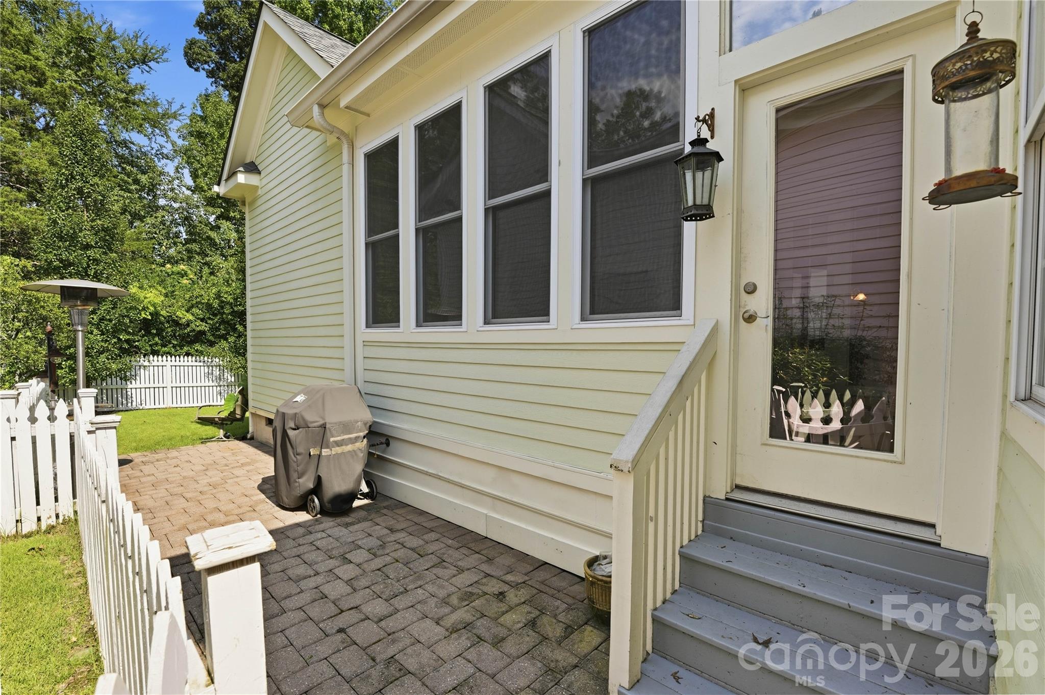 Paver patio beside a light-sided home, with gray steps, a glass-panel door, covered grill, and white picket fence.