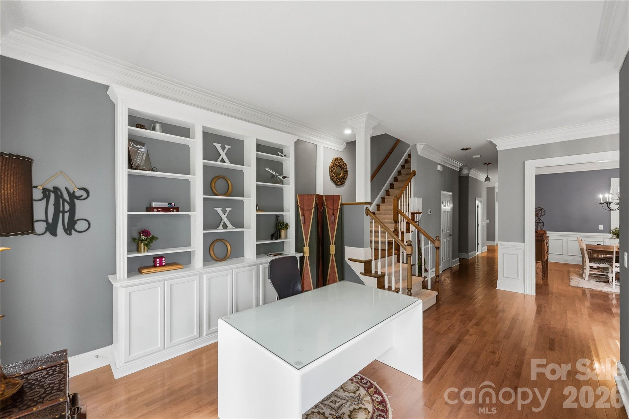 Office with white built-in shelving and glass-top desk beside a hardwood staircase.