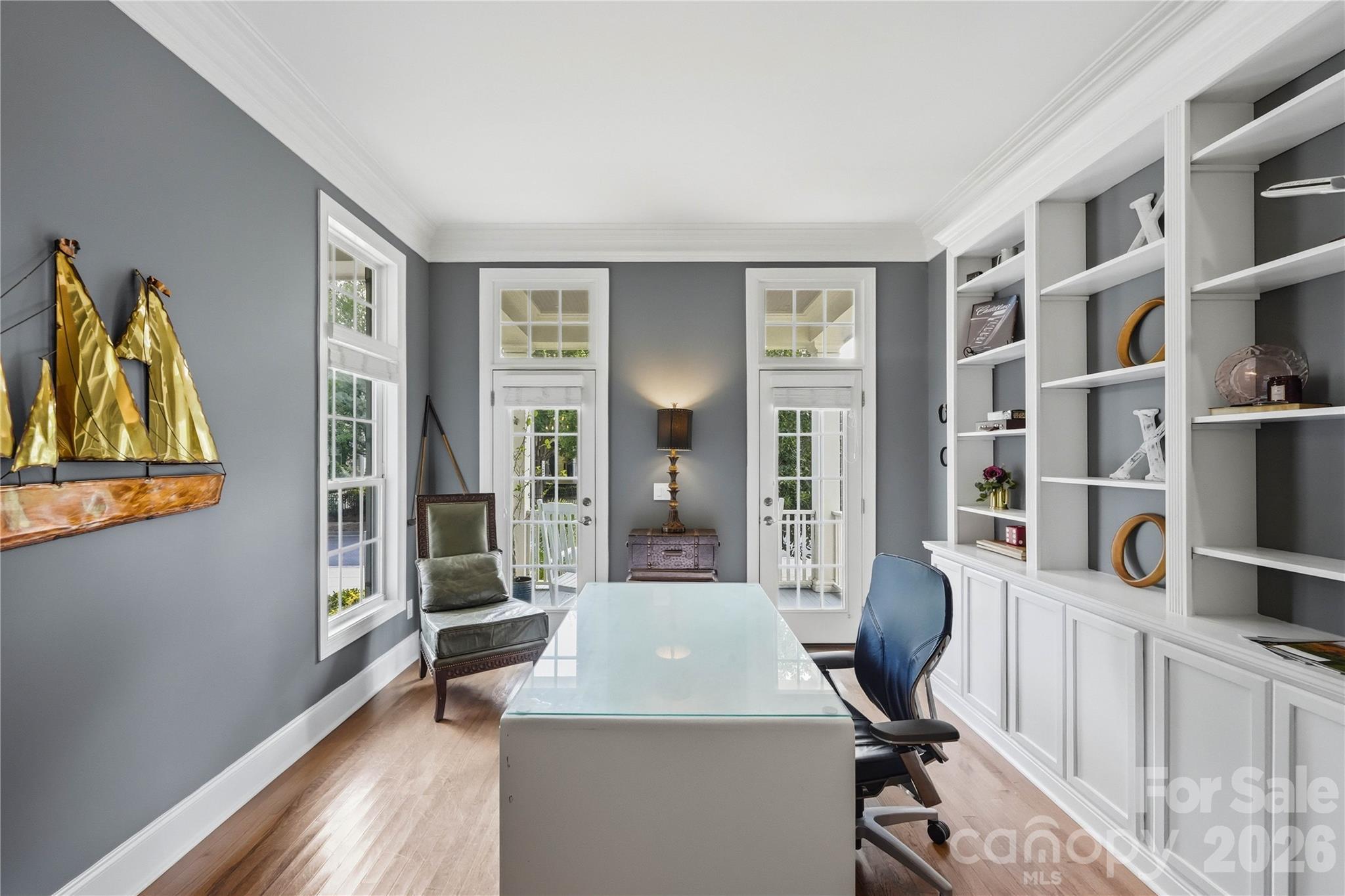 Furnished home office with white built-ins, glass desk, double glass doors, and gold sailboat wall art.