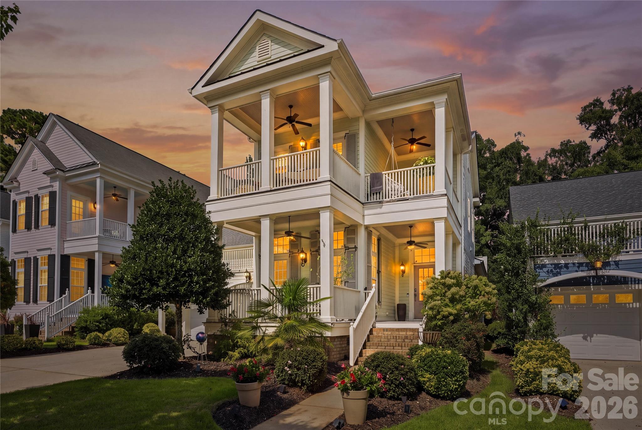 Two-story home with spacious covered porches, glowing windows, and landscaped gardens at dusk.