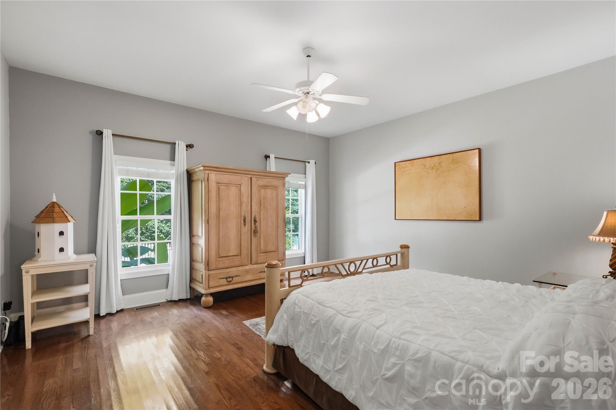Furnished bedroom with a white bed, wooden armoire, hardwood floors, and two curtained windows.