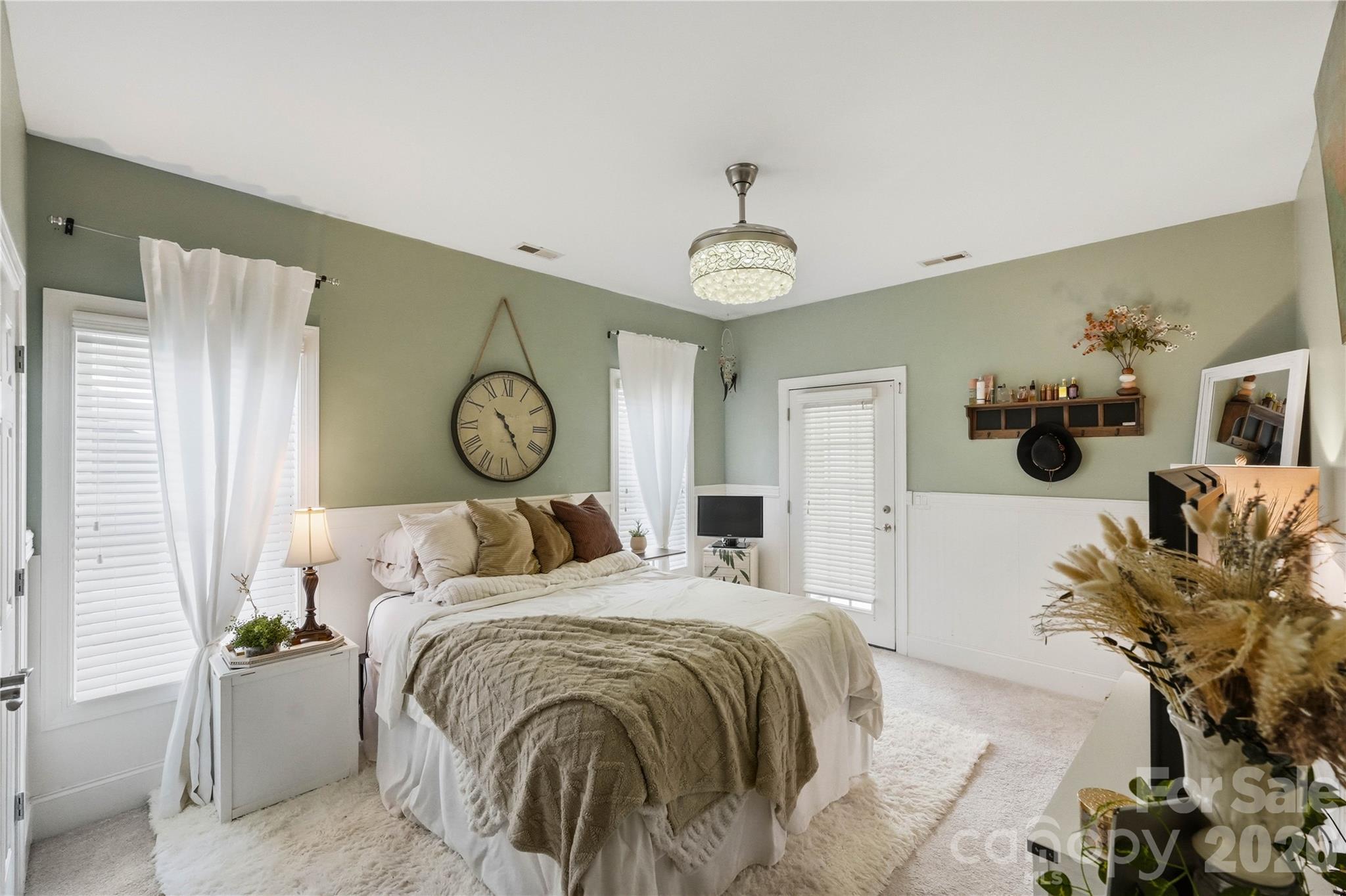 Furnished bedroom with sage-green walls, a layered cream bed, and a large hanging Roman-numeral clock.
