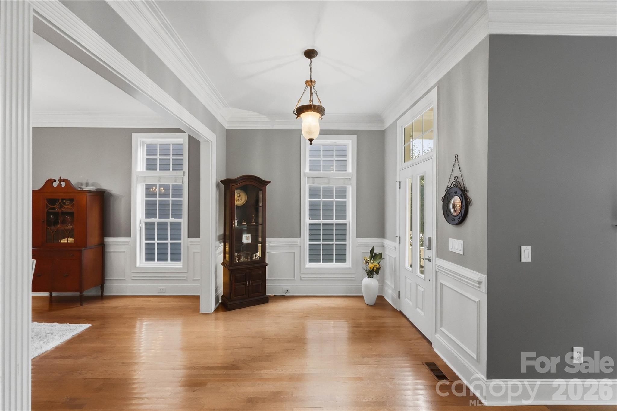 Furnished foyer with polished hardwood floors, paneled walls, antique cabinets, tall windows, and a glass-paneled front door.