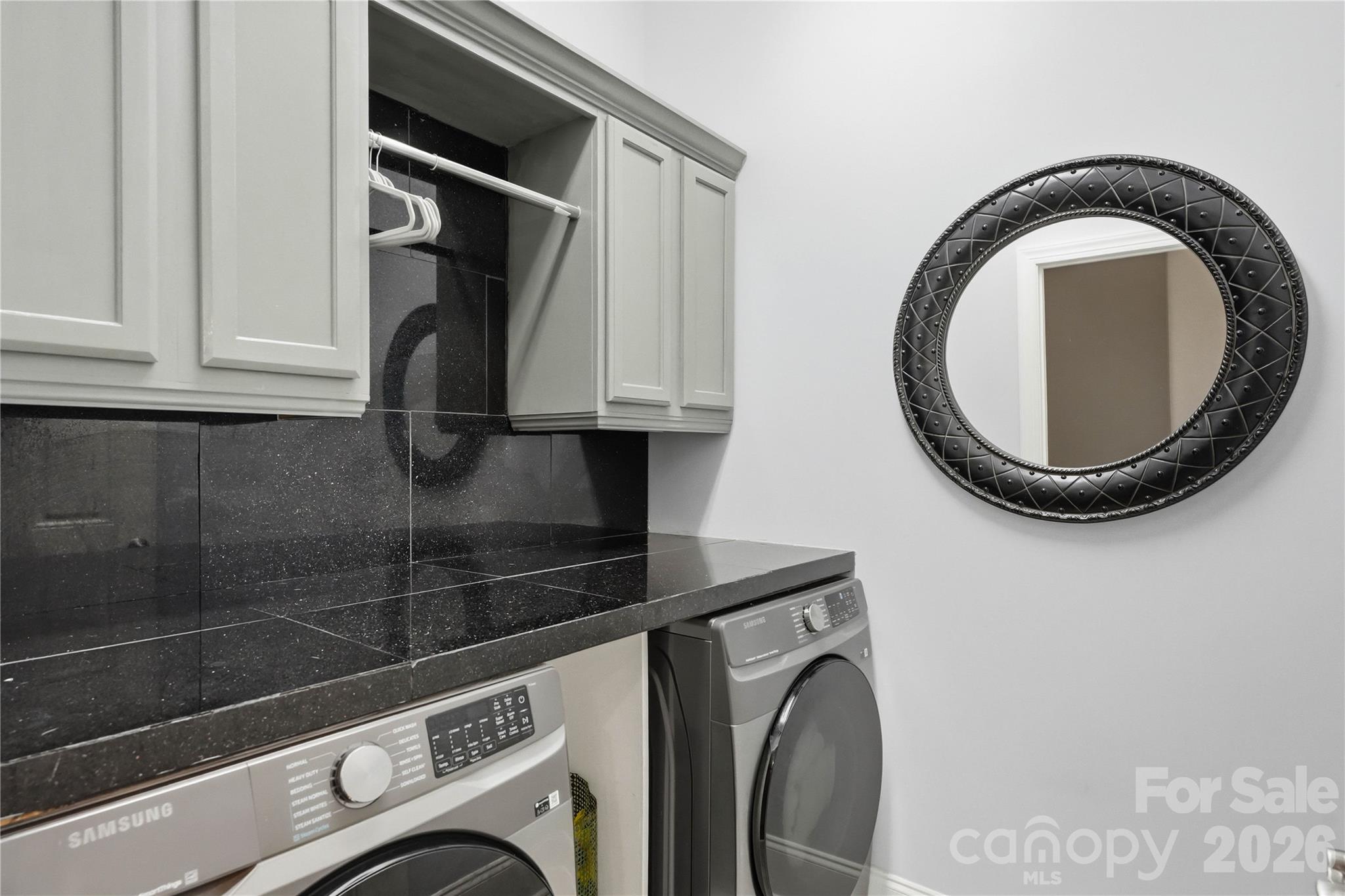 Laundry room with front-loading washer and dryer, black tile counter, white cabinets, and round mirror.