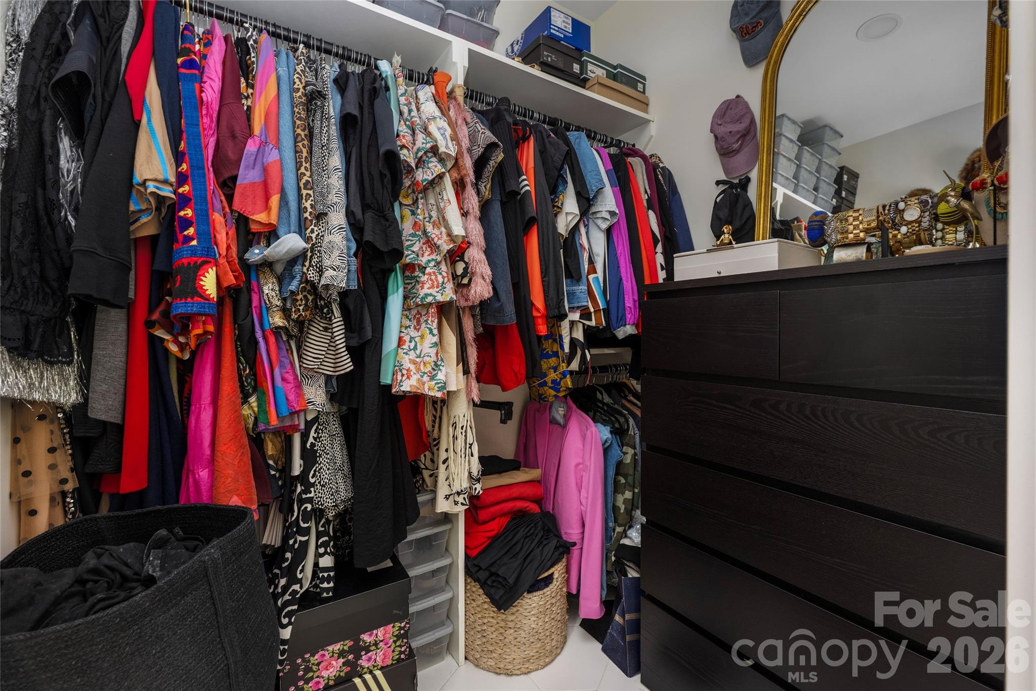 Walk-in closet with colorful clothing racks, storage baskets, and a dark dresser with a gold-framed mirror.