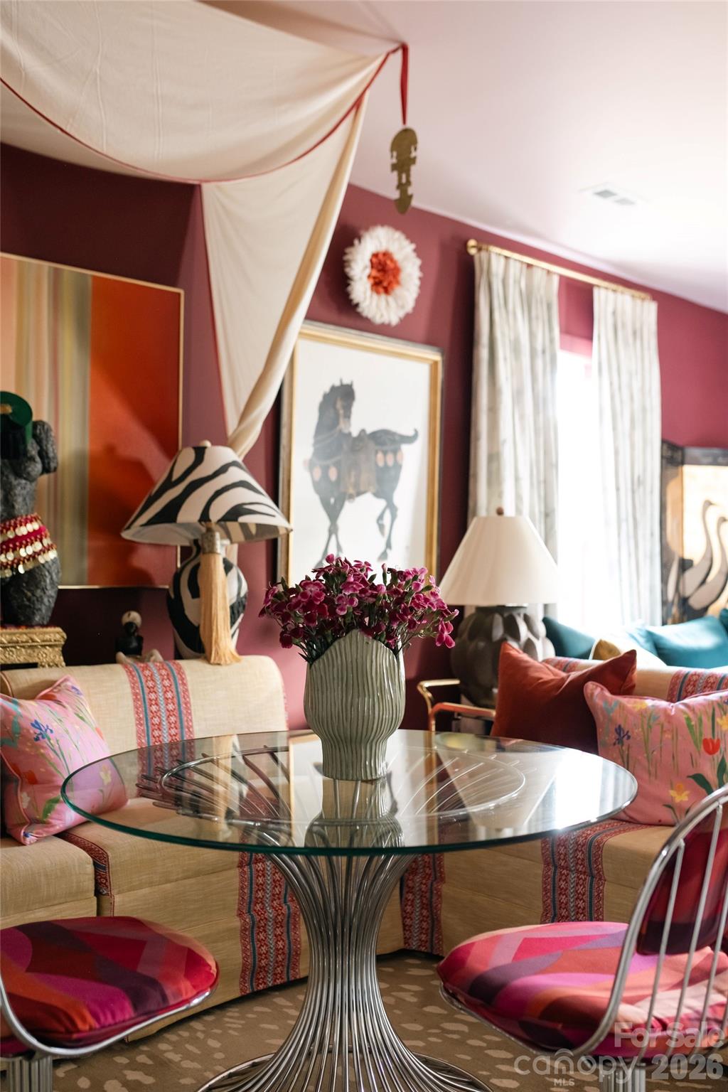 Dining nook with a glass table, cream banquette, patterned pink cushions, and burgundy walls.