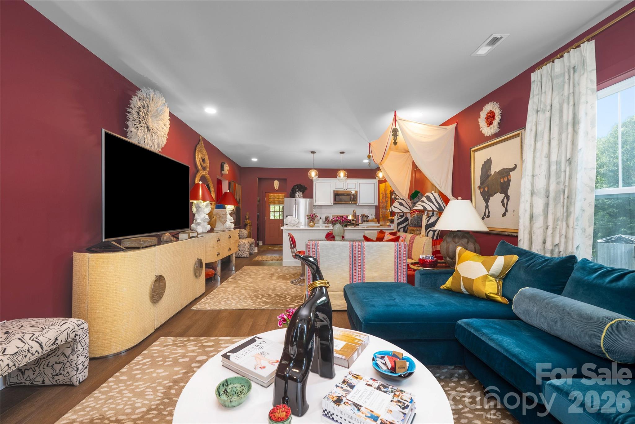 Open-plan living room with burgundy walls, teal sectional, large television, and kitchen beyond.