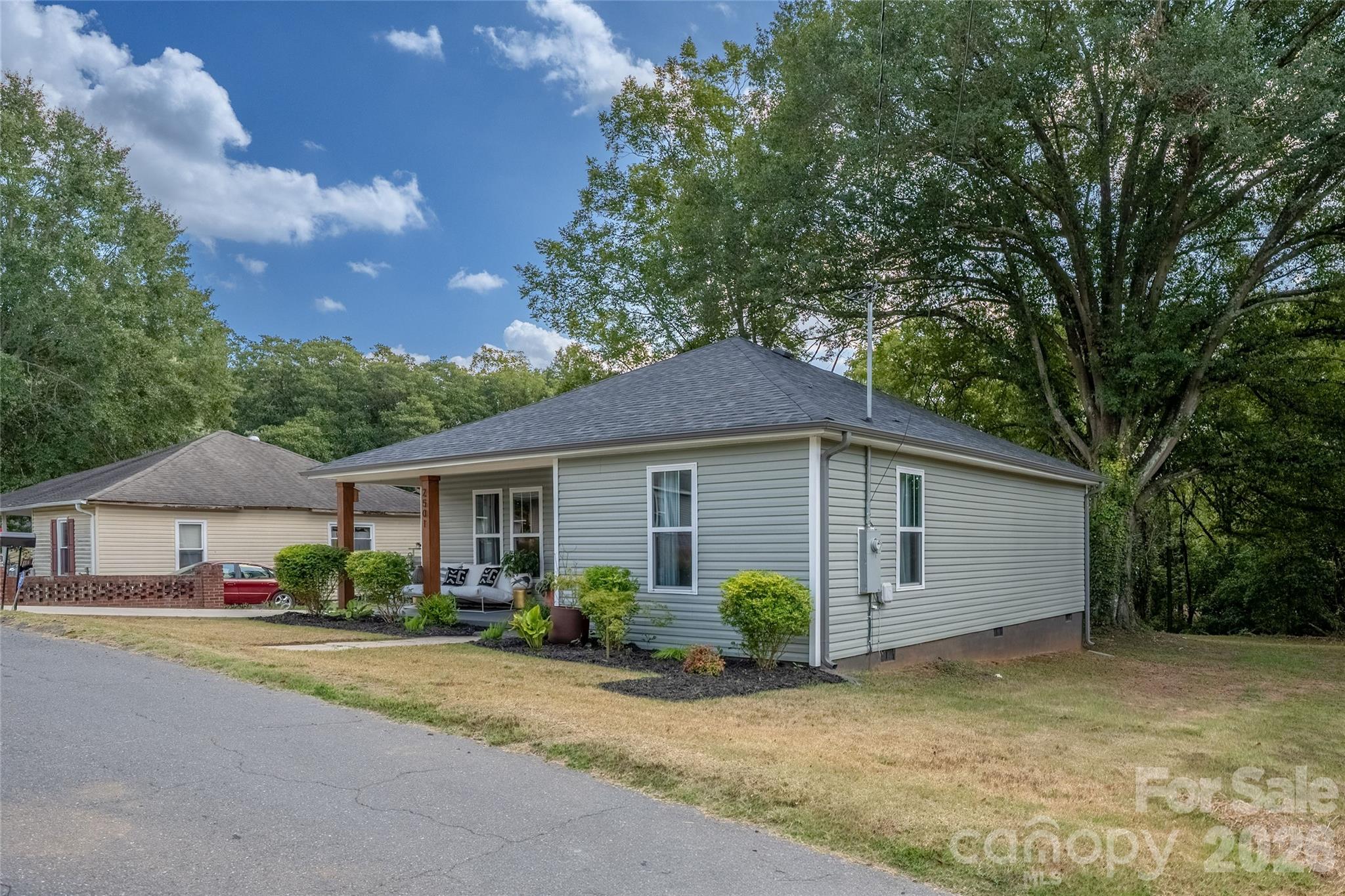 Gray single-story house with a covered porch, landscaped shrubs, and mature trees.