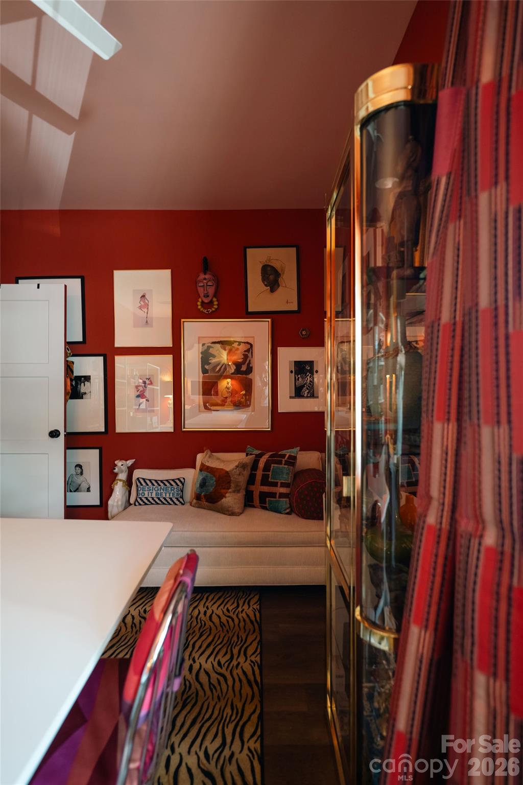 Office nook with a white desk, cream sofa, red art-covered walls, glass display cabinet, and patterned rug.