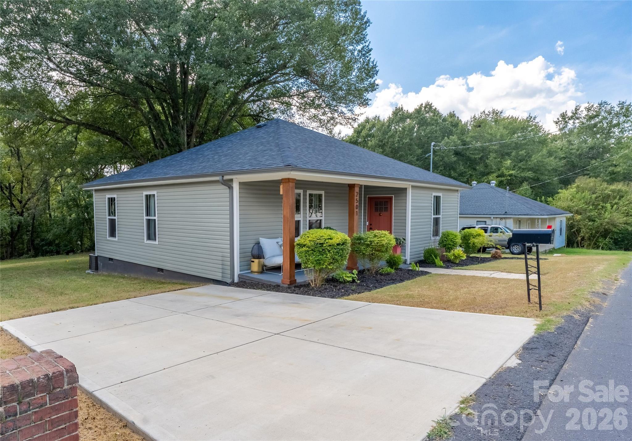 Gray single-story home with a covered porch, red door, landscaped shrubs, and wide concrete driveway.