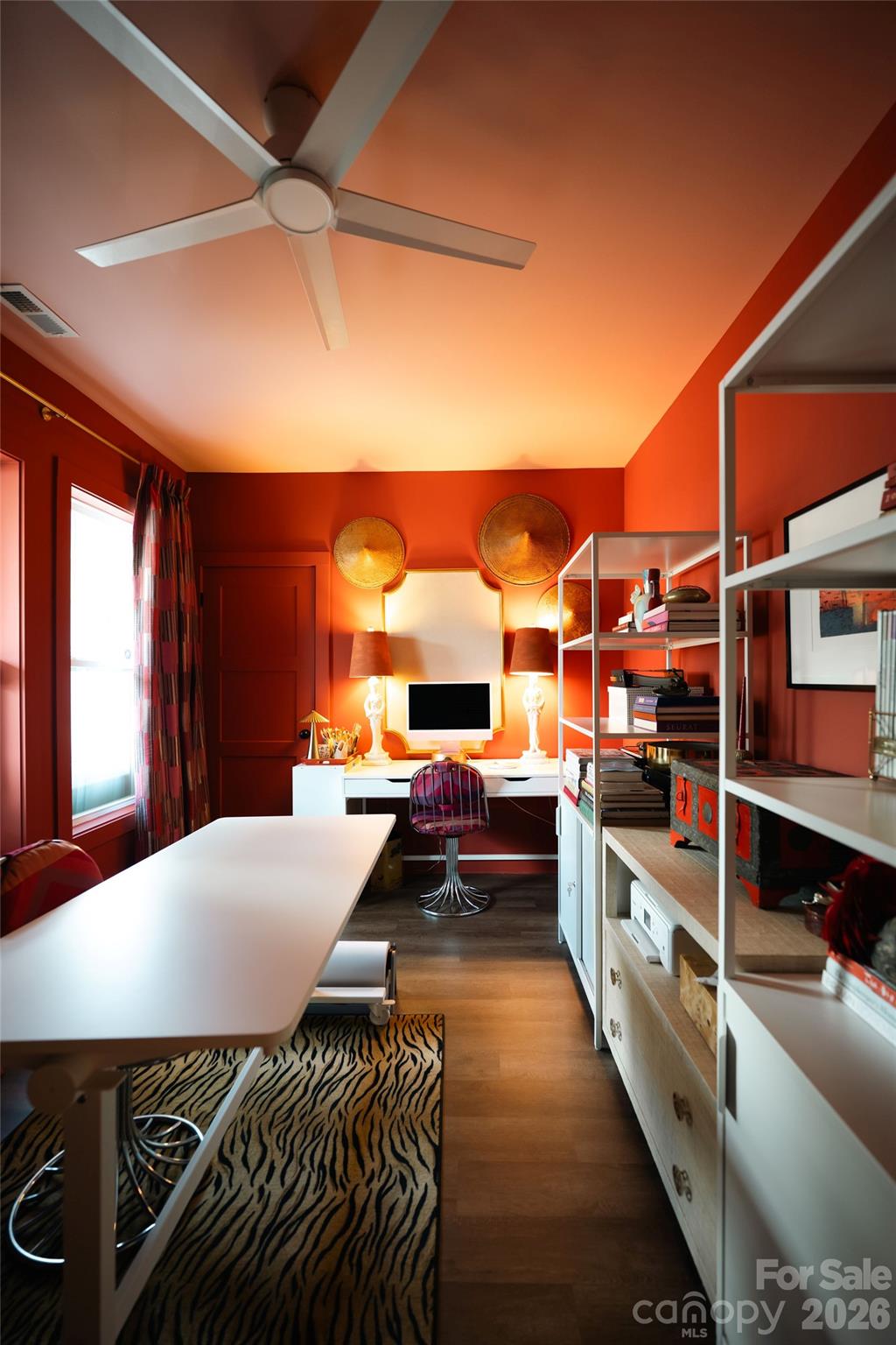 Home office with vivid red-orange walls, white desk, open shelving, and zebra-patterned rug.