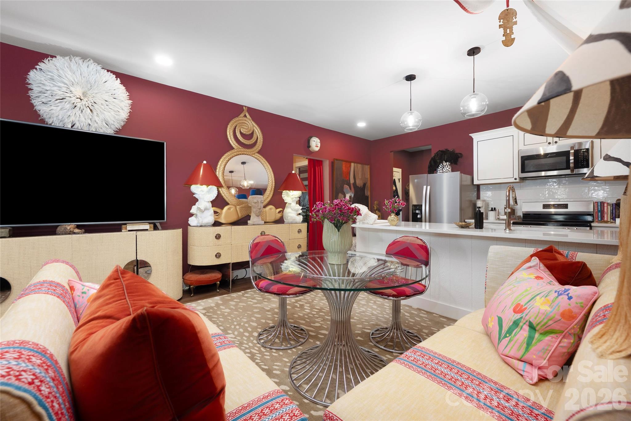Open-plan living and dining area with burgundy walls, colorful sofas, glass table, and adjoining white kitchen.