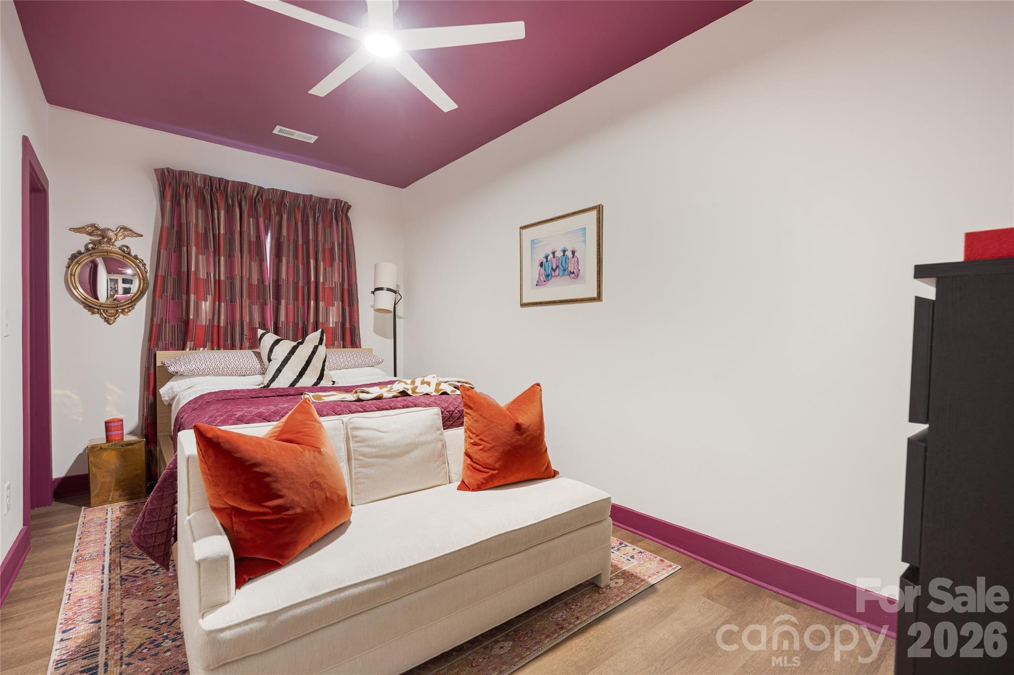 Staged bedroom with a cream bench, orange pillows, patterned curtains, and burgundy ceiling.
