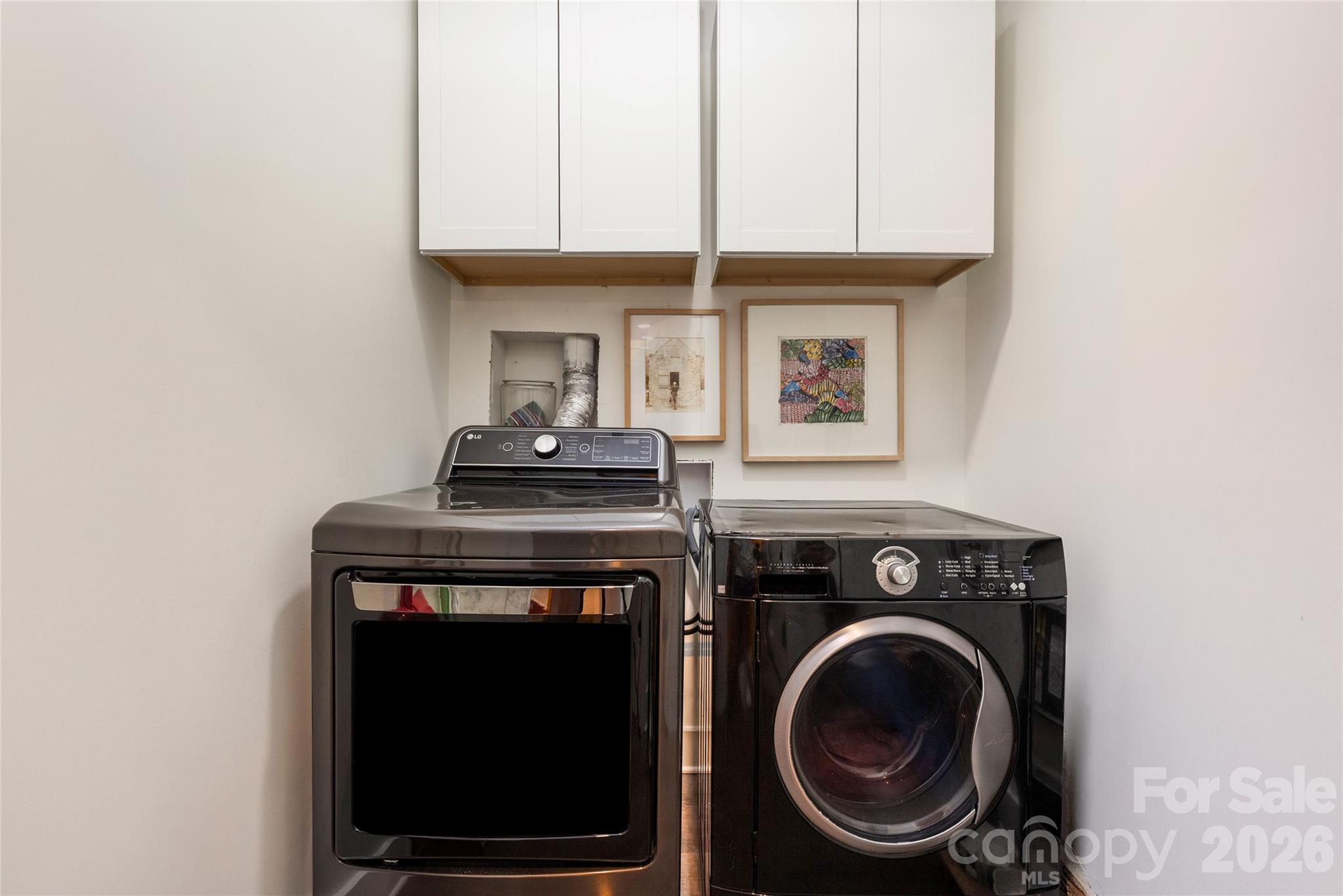 Laundry room with dark washer and dryer beneath white cabinets and framed artwork.