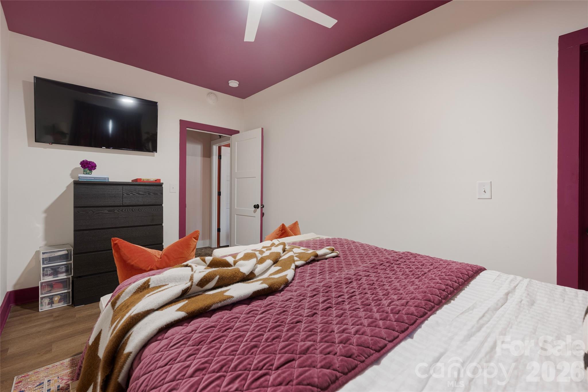 Furnished bedroom with a magenta quilted bedspread, orange pillows, and a wall-mounted television.