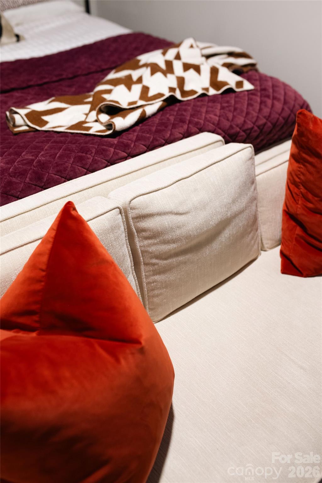 Bedroom with a burgundy quilted bedspread, cream upholstery, and bright orange-red pillows.