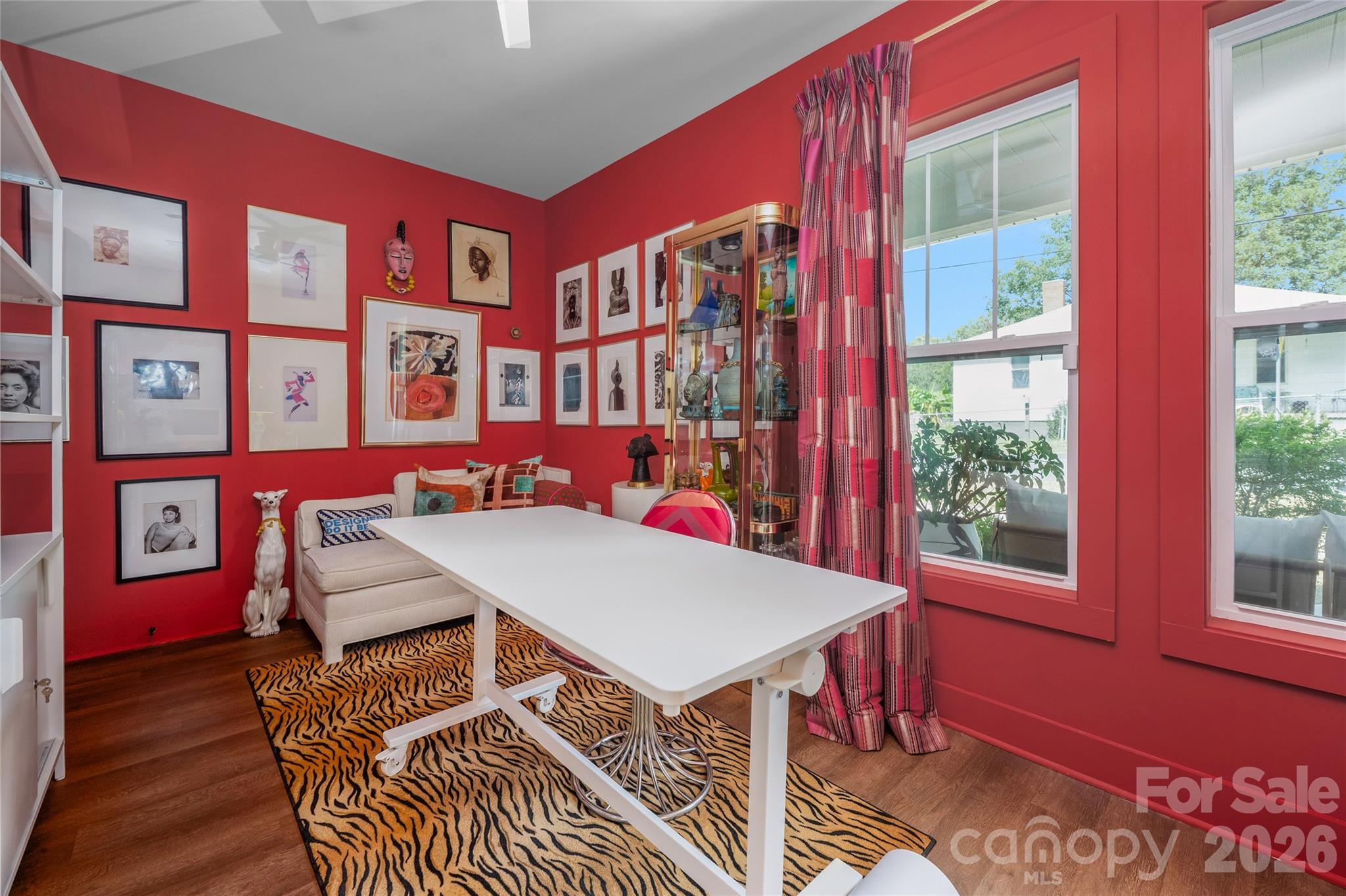 Red-walled home office with a white desk, gallery wall, glass cabinet, and tiger-stripe rug.
