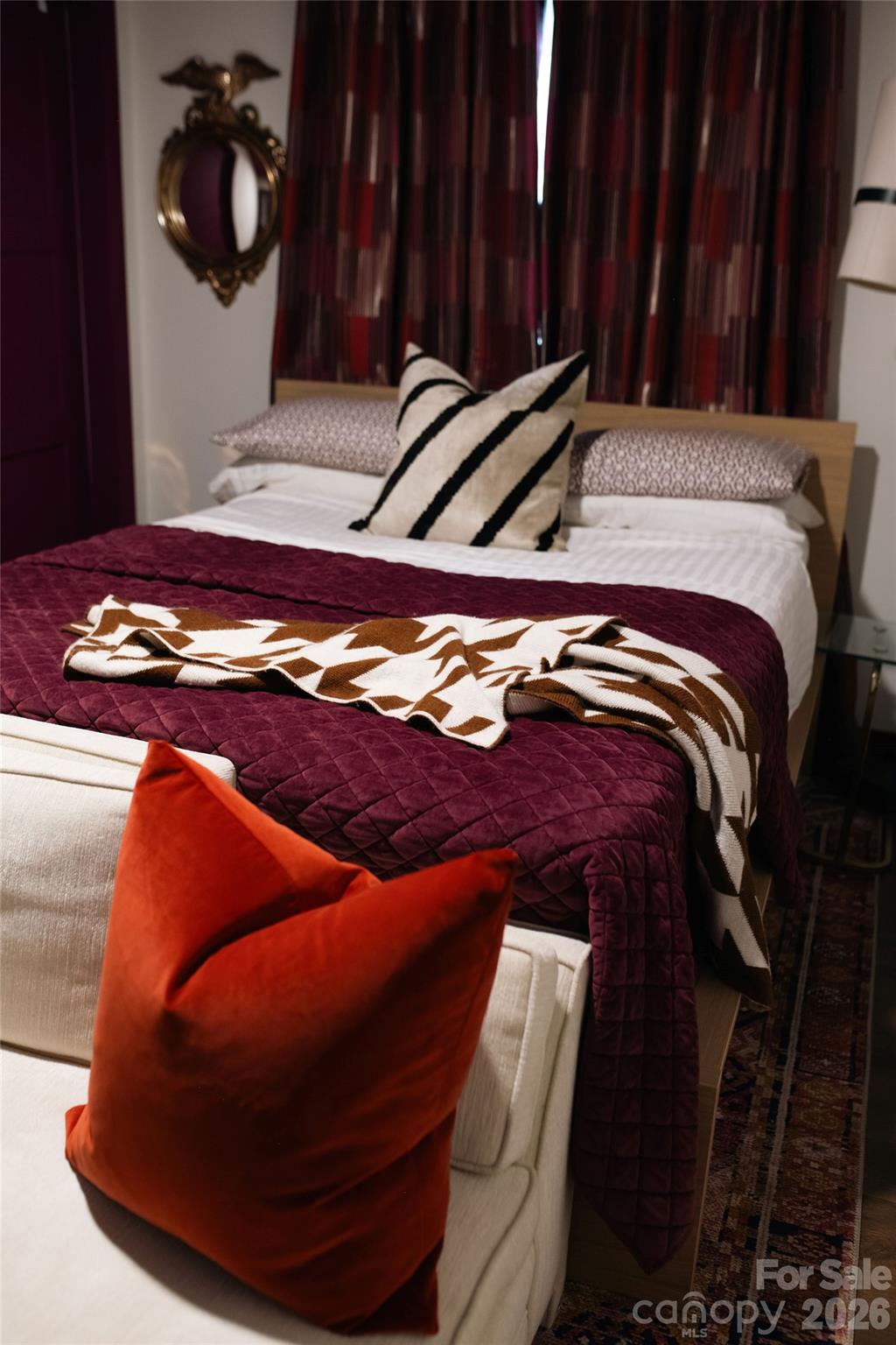 Staged bedroom with a burgundy quilt, patterned pillows, orange cushion, and ornate wall mirror.