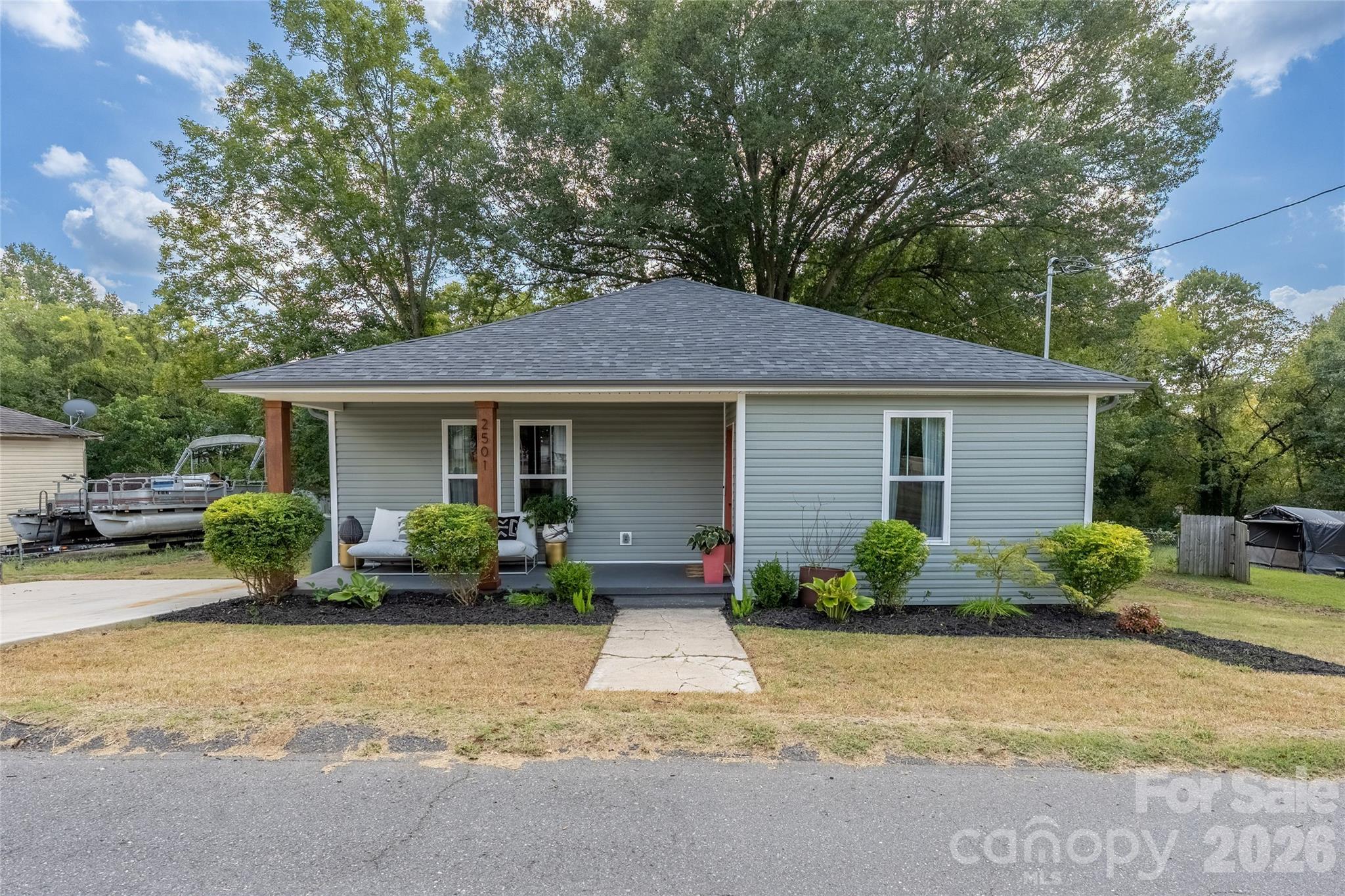 Single-story gray house with a covered porch, landscaped shrubs, and mature trees behind it.