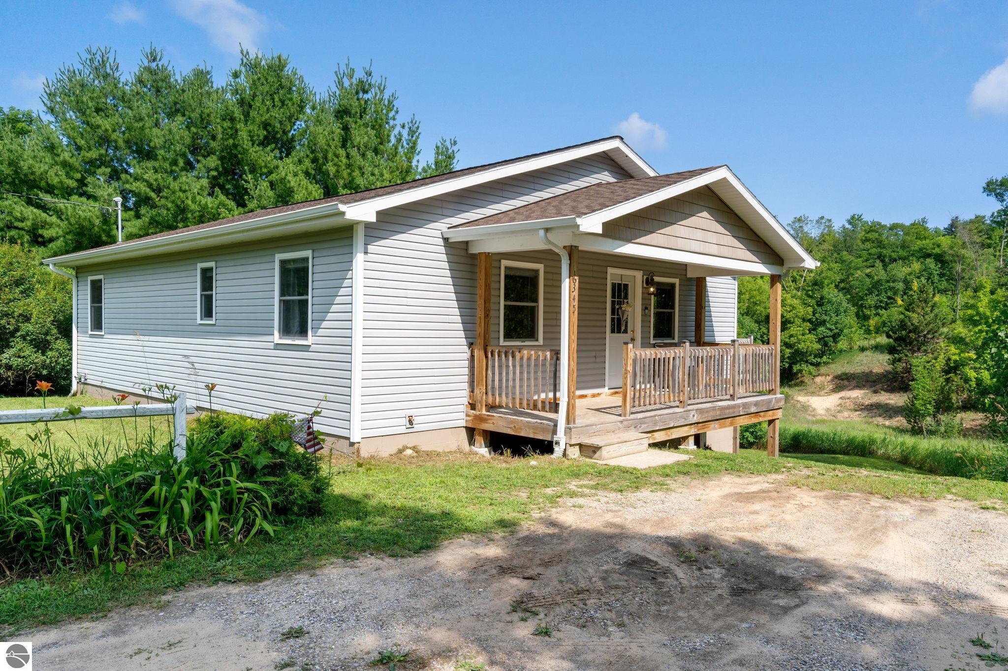 Gray single-story home with a covered wooden porch, gravel driveway, and leafy surroundings.