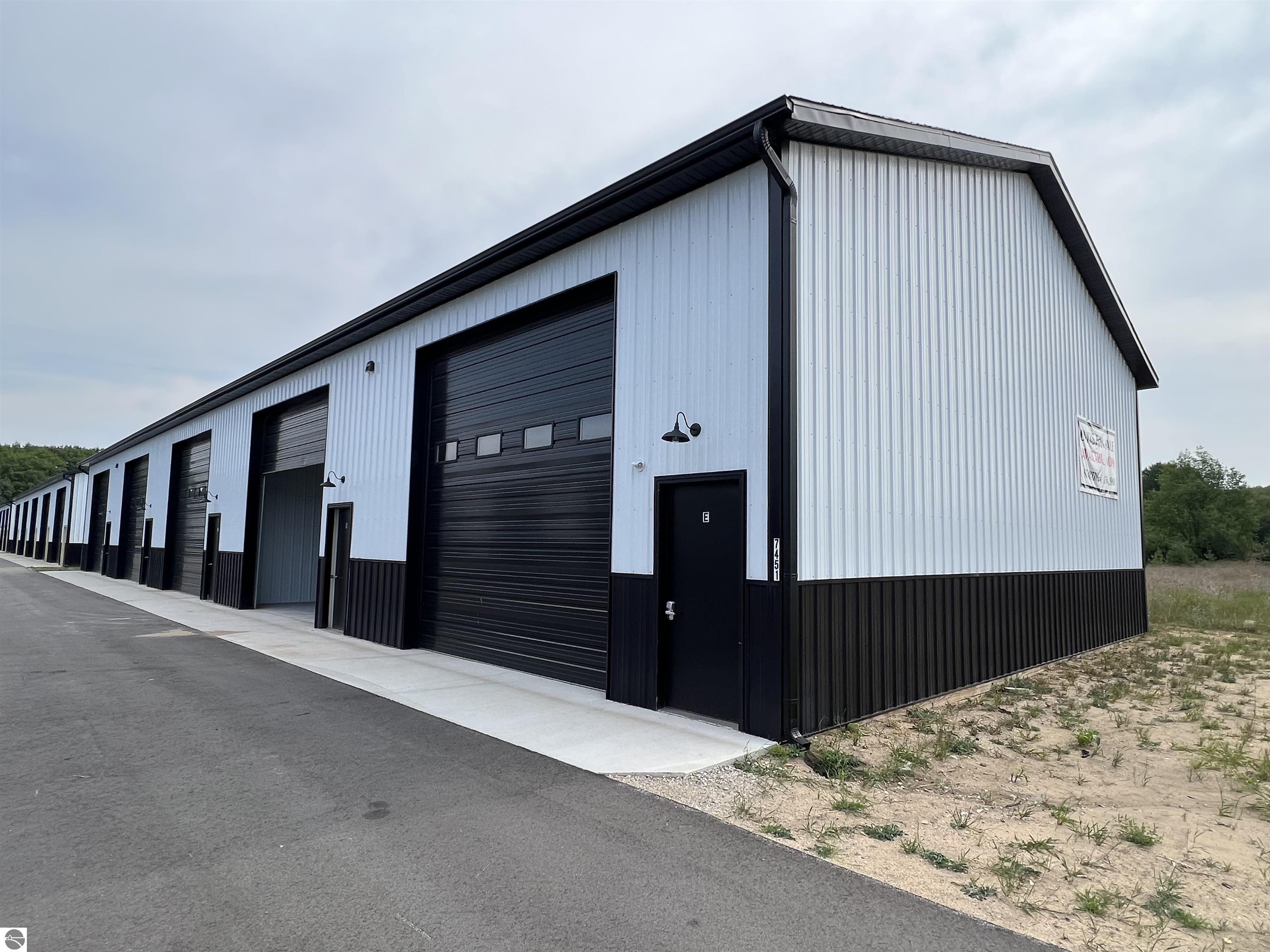 Long white-and-black metal commercial building with multiple garage doors beside a paved lane.