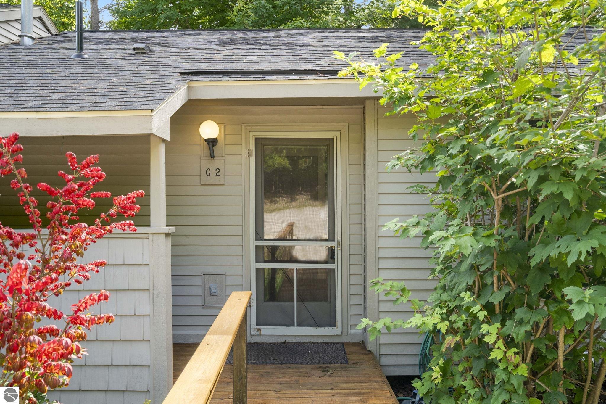 Gray-sided home entrance with screened door marked G 2, wooden porch railing, and surrounding foliage.