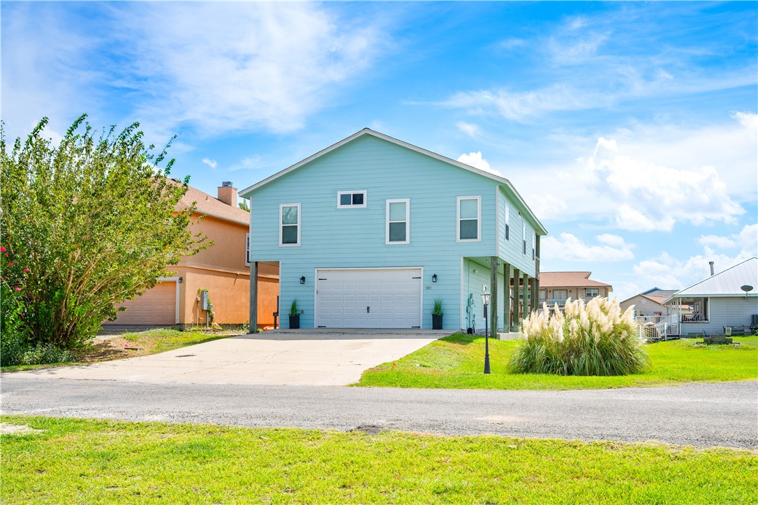 Light blue raised two-story house with a garage, broad driveway, and ornamental grass.