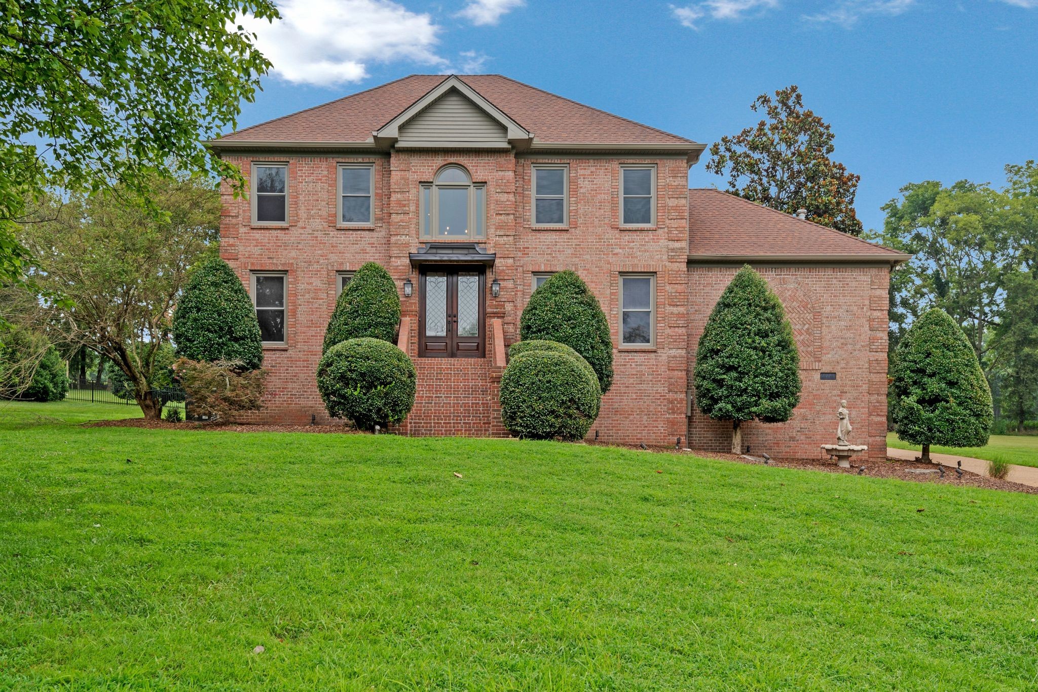 Two-story brick house with sculpted shrubs, a broad lawn, and a small statue fountain.