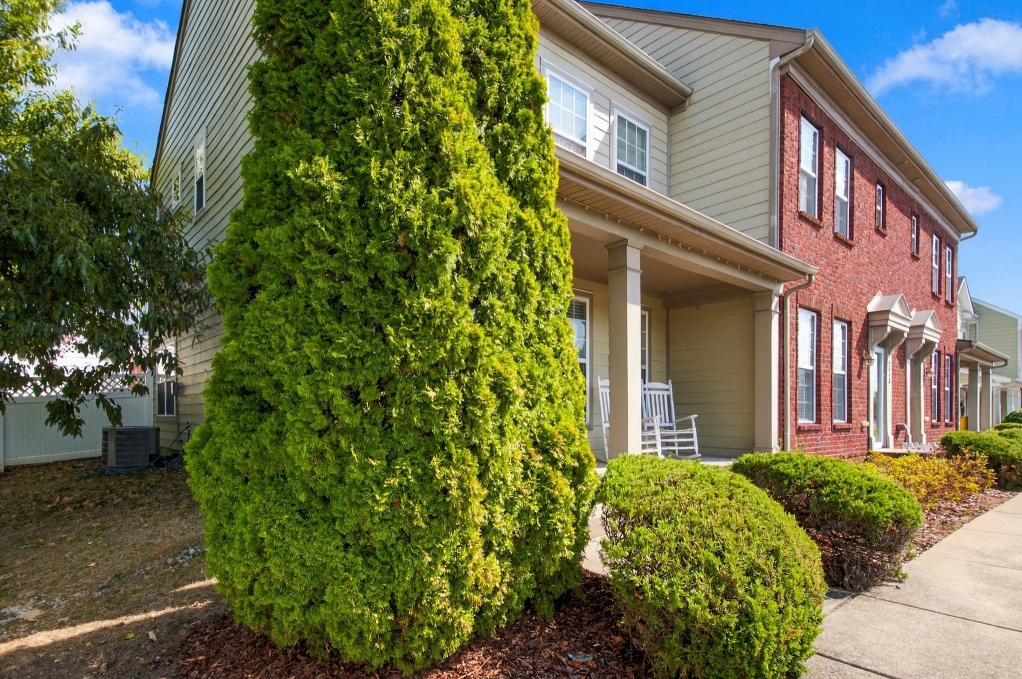 Two-story townhouse with red brick, beige siding, a covered porch, and landscaped shrubs.