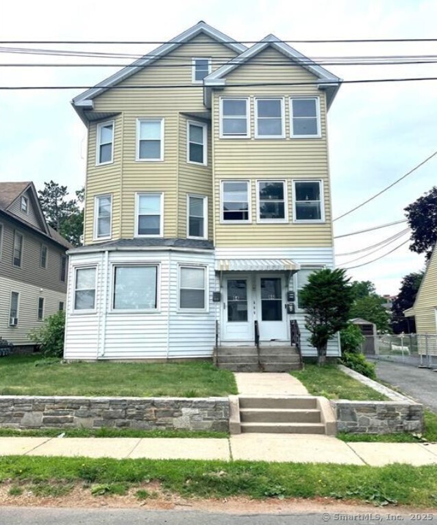 Three-story yellow-sided multifamily building with white trim, covered entrance, and stone retaining wall.