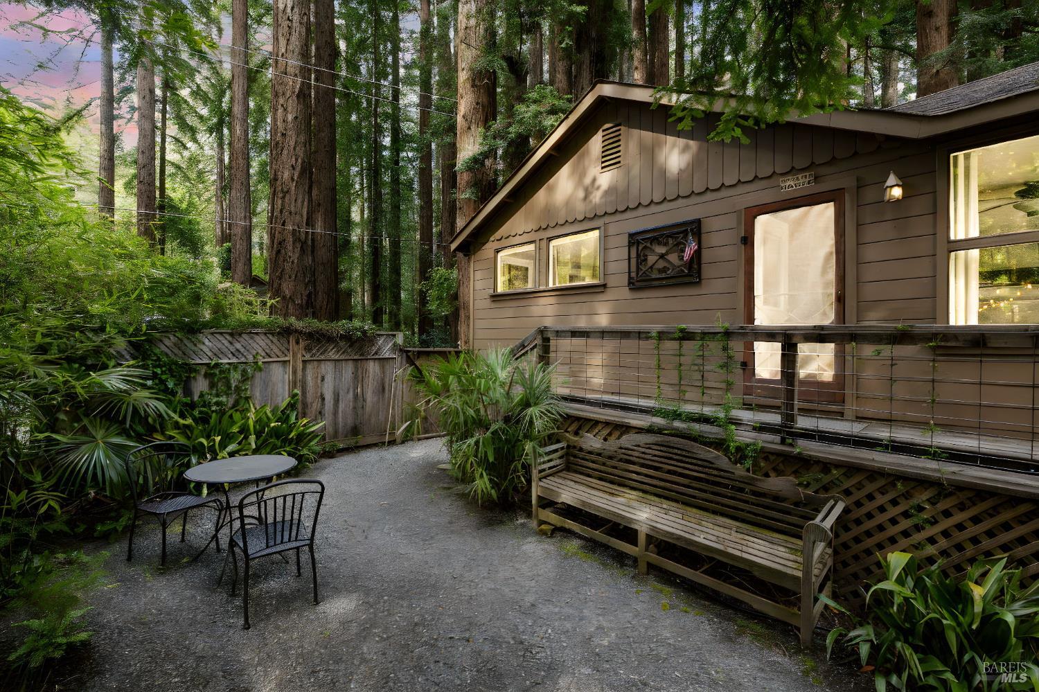 Brown wood-sided home nestled among redwoods, with a gravel patio and raised deck seating.