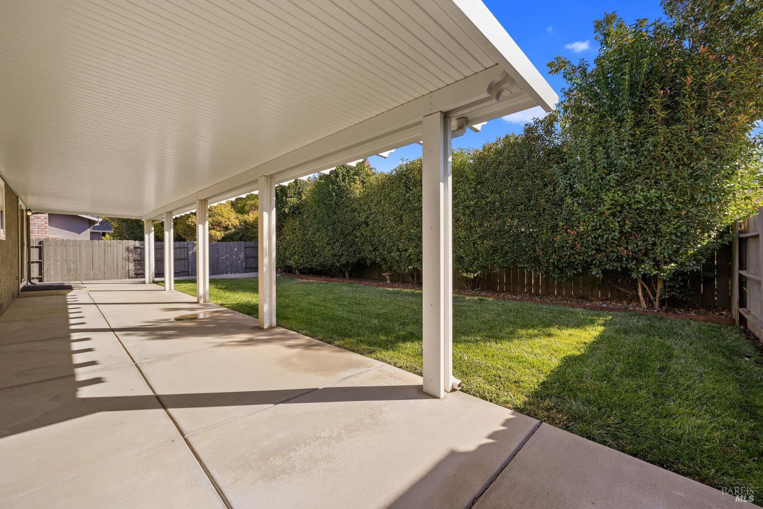 Covered patio beside a fenced lawn, with white posts and tall privacy hedges.