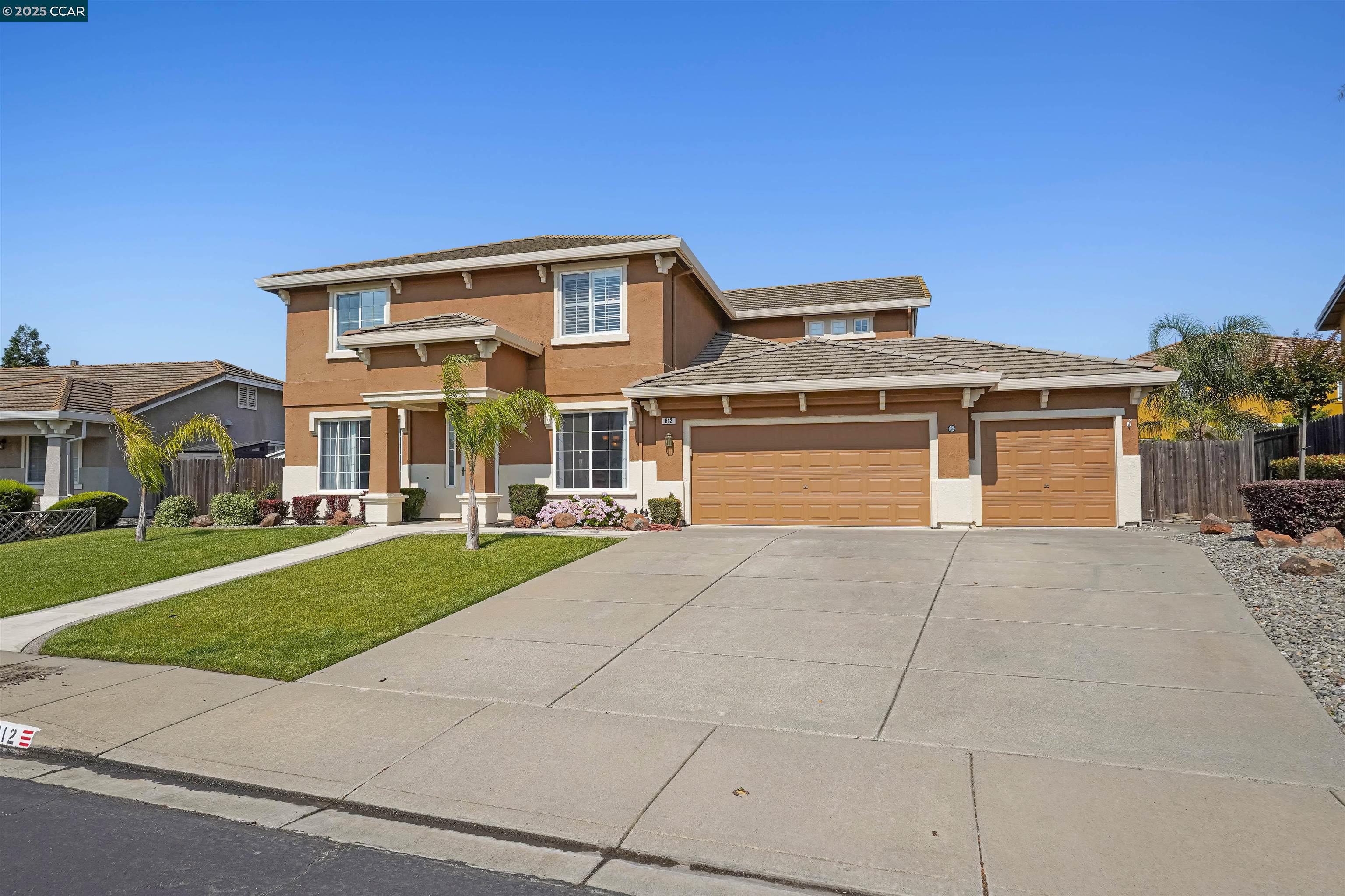 Two-story tan stucco home with a three-car garage, broad driveway, and landscaped front lawn.