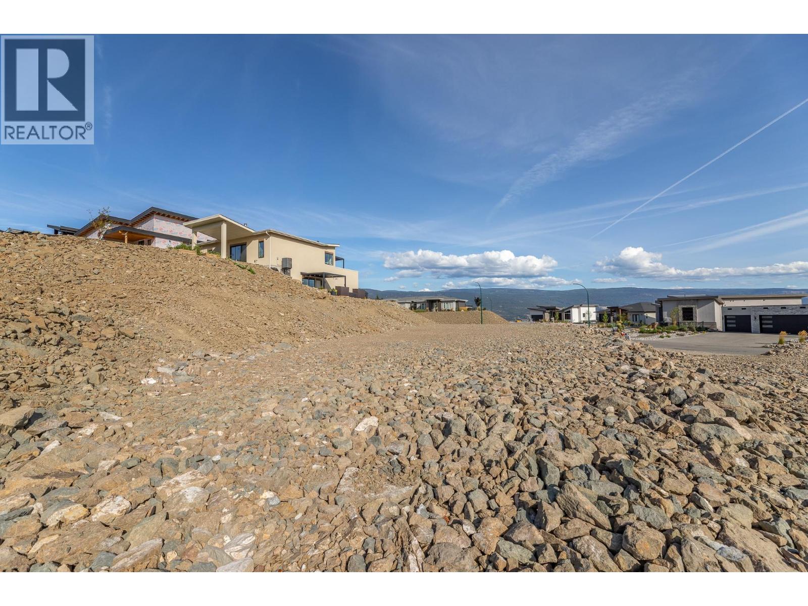 Rocky sloping lot beside modern homes, with distant hills and a wide blue sky.