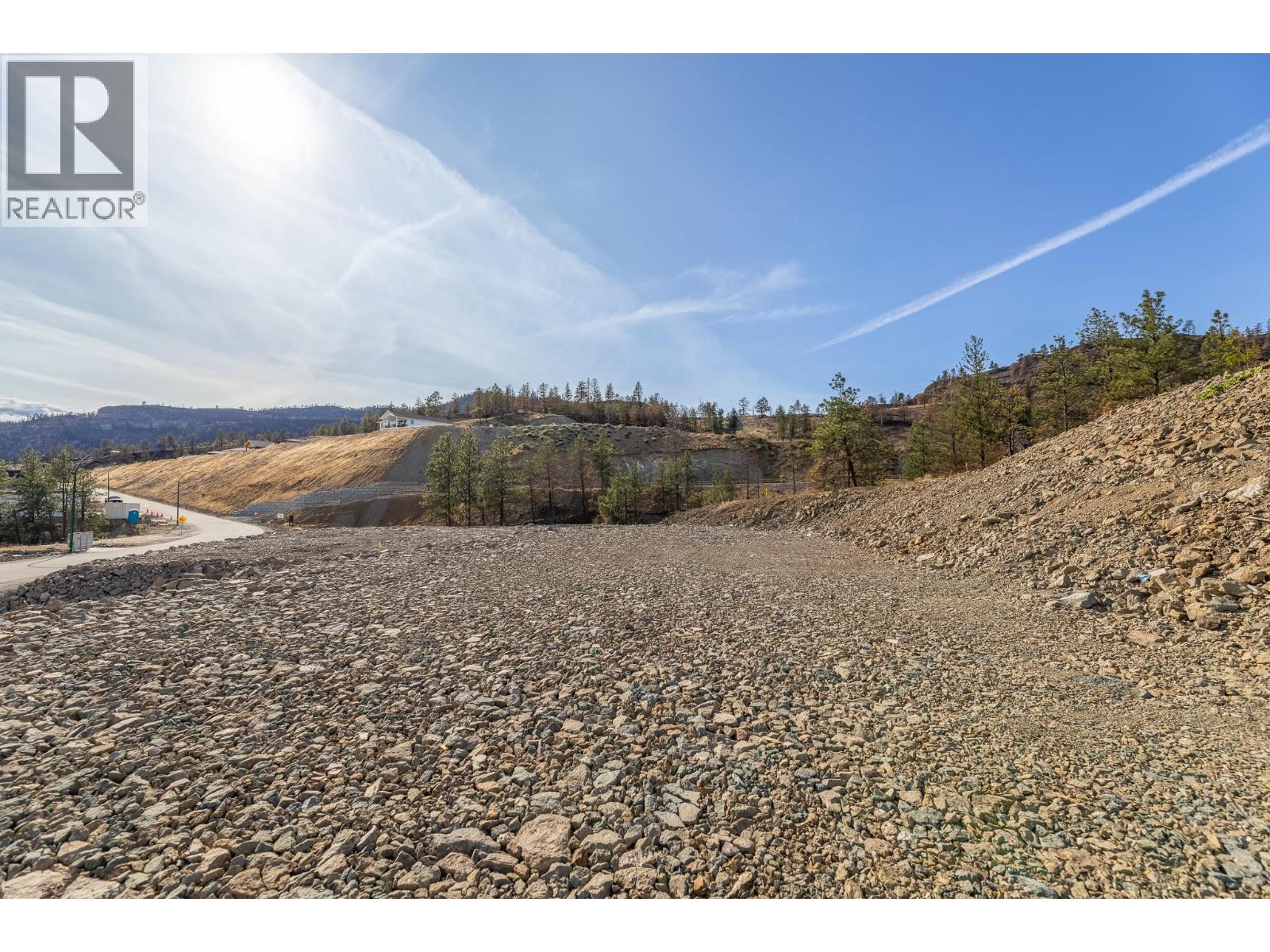 Rocky graded lot on a hillside, with a road and sparse trees nearby.