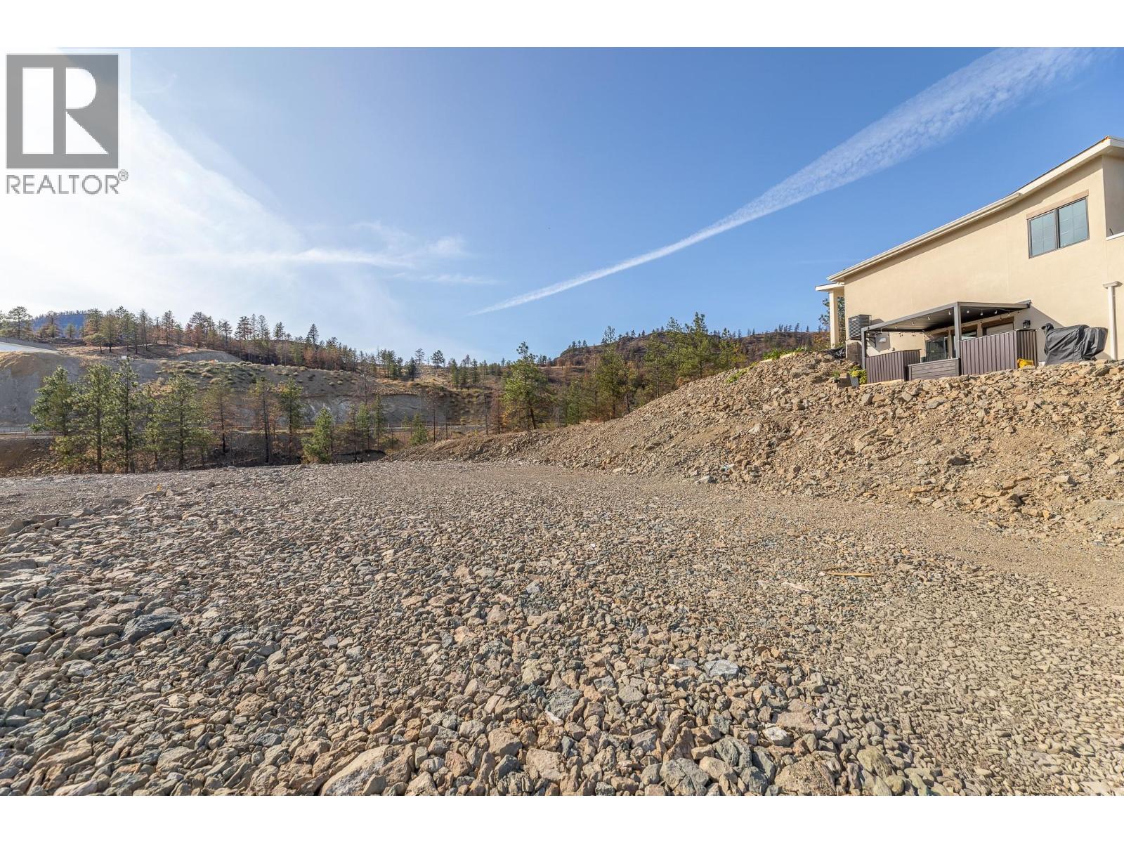 Rocky vacant lot beside a beige house, with sparse trees and dry hills beyond.