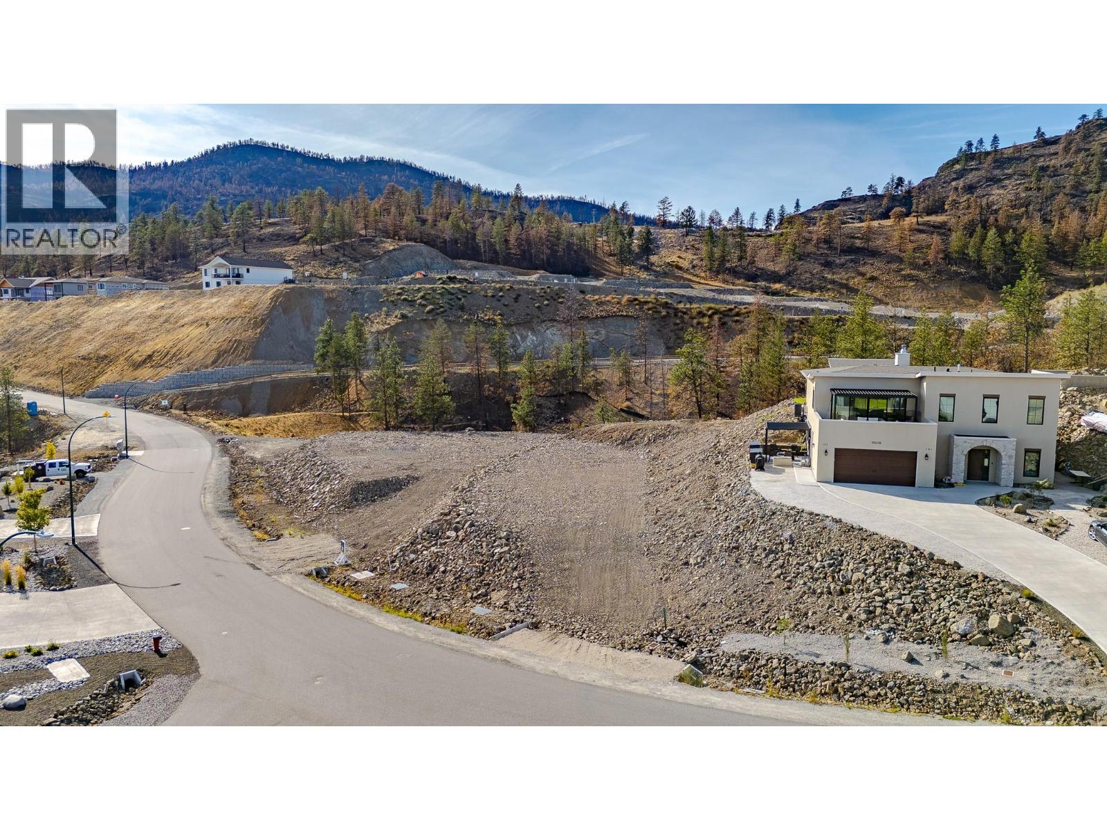 Vacant hillside lot beside a curving road, with nearby homes and wooded mountains beyond.