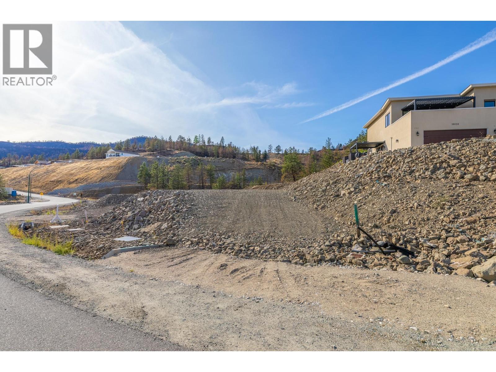 Vacant rocky lot beside a road, with a modern house on the slope and dry hills beyond.