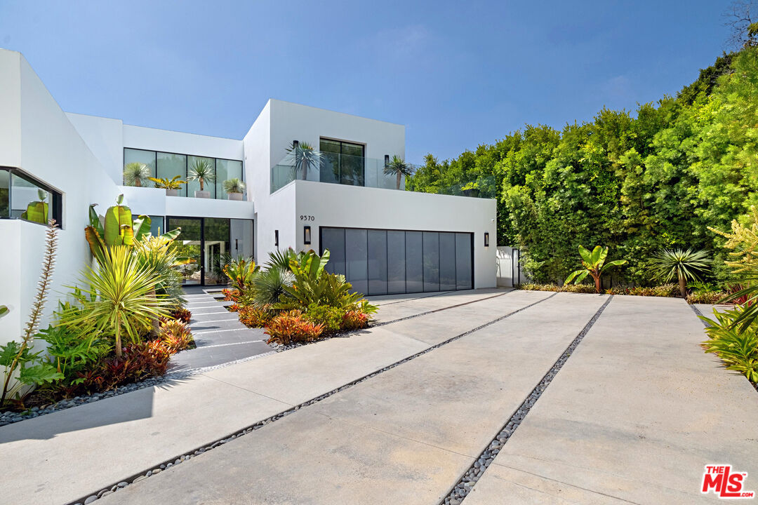 Modern white home with tropical landscaping, dark garage doors, and a broad concrete driveway.
