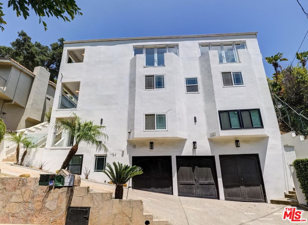 White multi-story home with glass balconies, three black garage doors, and a sloped driveway.