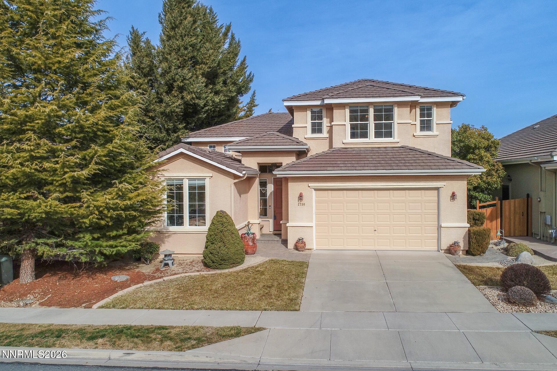 Two-story beige stucco house with a double garage, tiled roof, and mature evergreens.