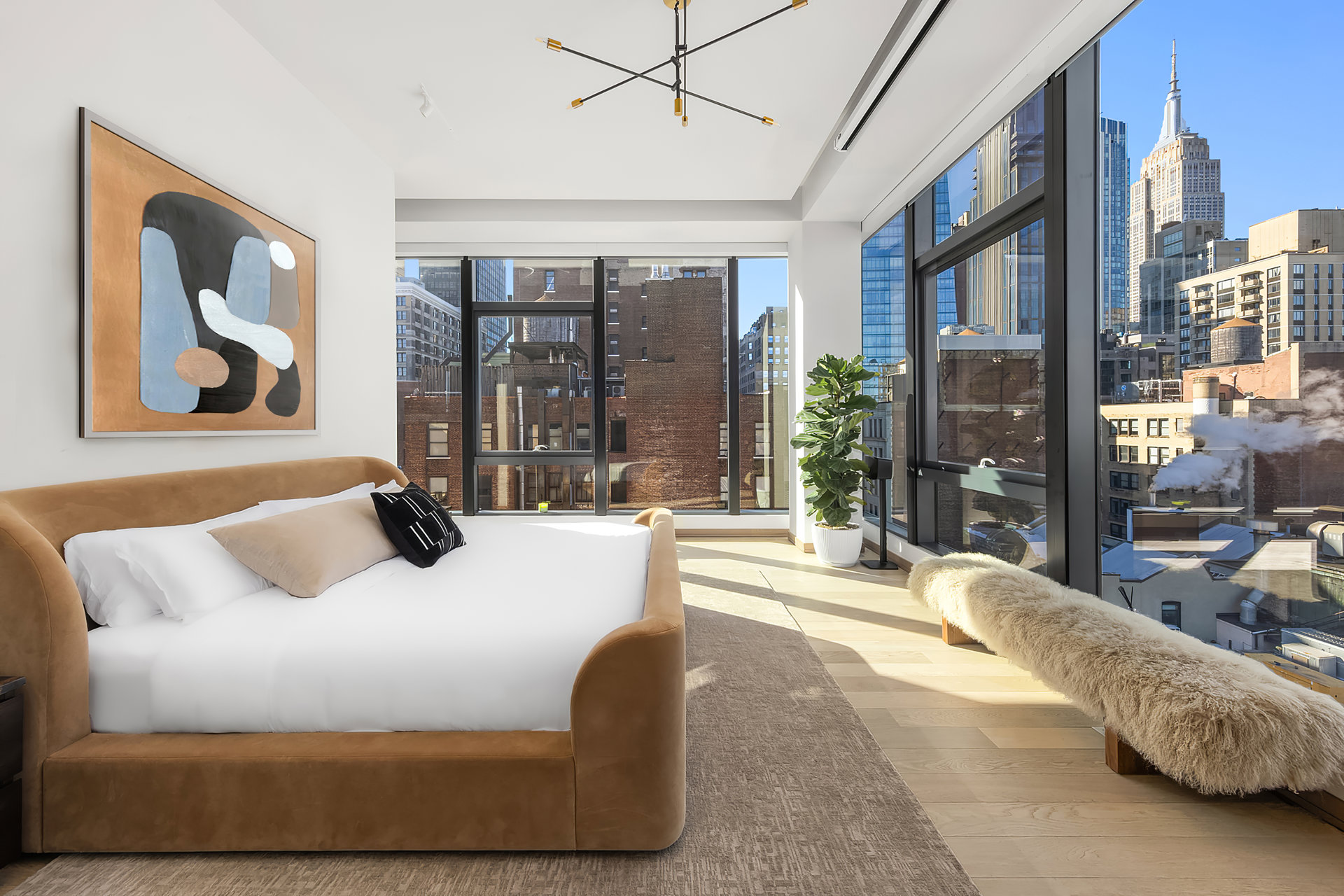 Staged bedroom with a tan bed and floor-to-ceiling windows overlooking the Empire State Building.