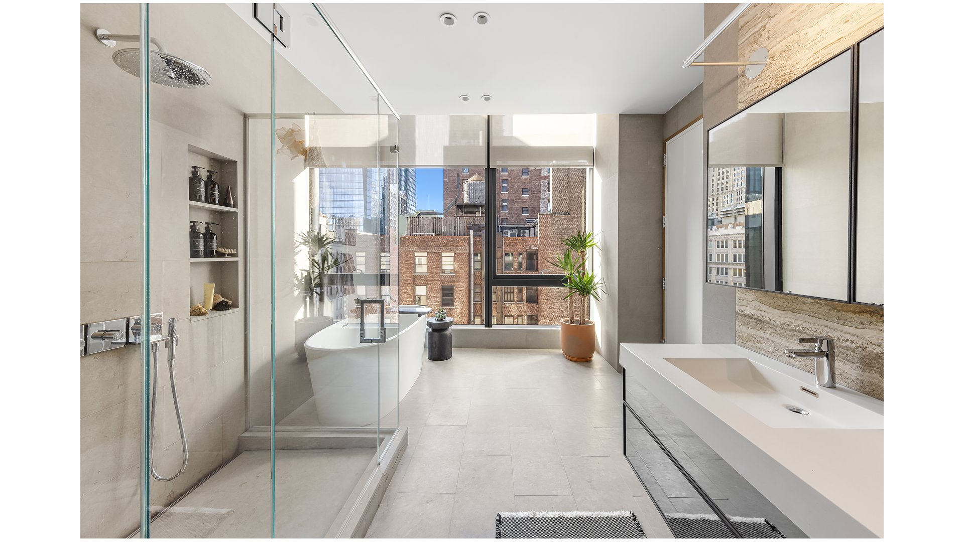 Modern bathroom with a glass shower, freestanding tub, and large windows overlooking city buildings.