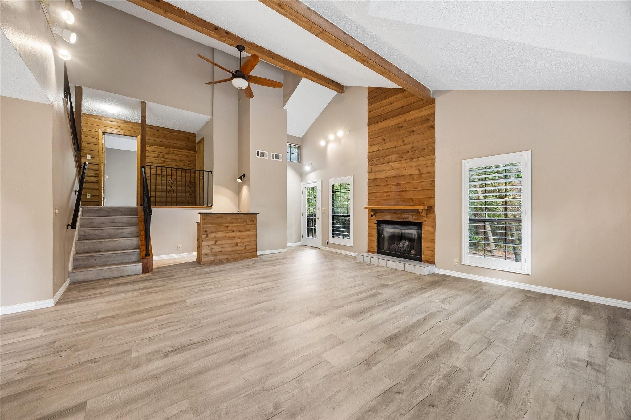 Empty living room with a wood-clad fireplace, vaulted beam ceiling, and staircase.