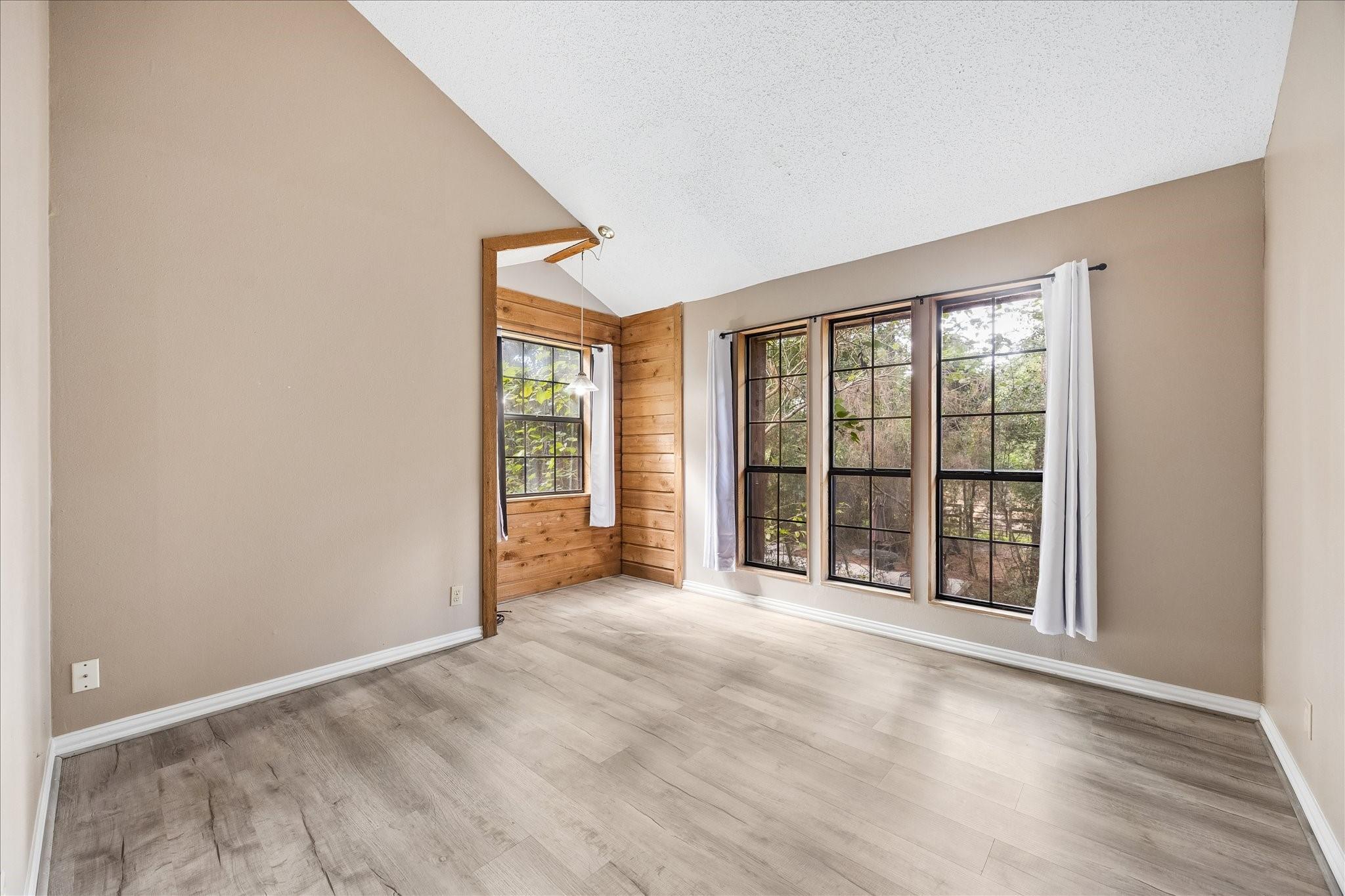 Vacant room with a vaulted ceiling, wood-paneled alcove, and large windows overlooking trees.